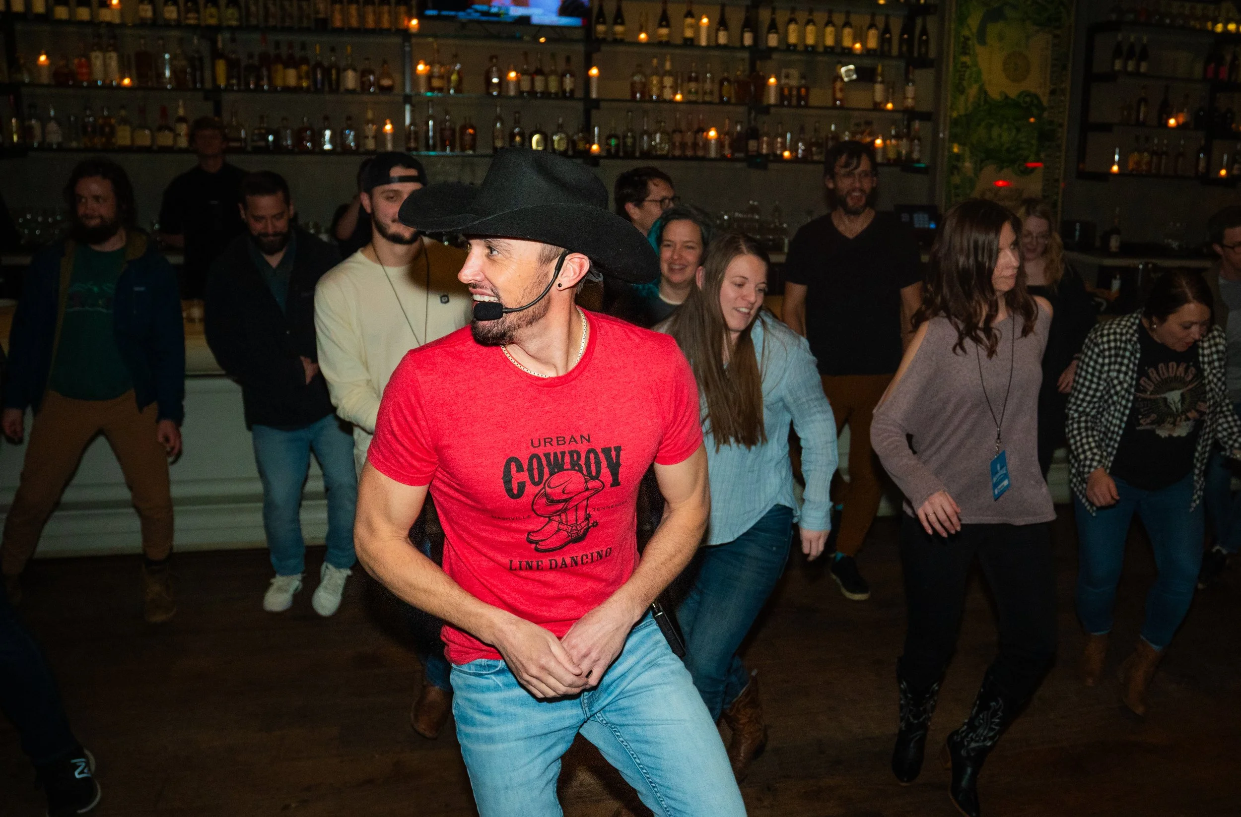 A group of people line dancing in a dimly lit bar with shelves of bottles and candles in the background. The man in the foreground wears a black cowboy hat, red shirt, and jeans.