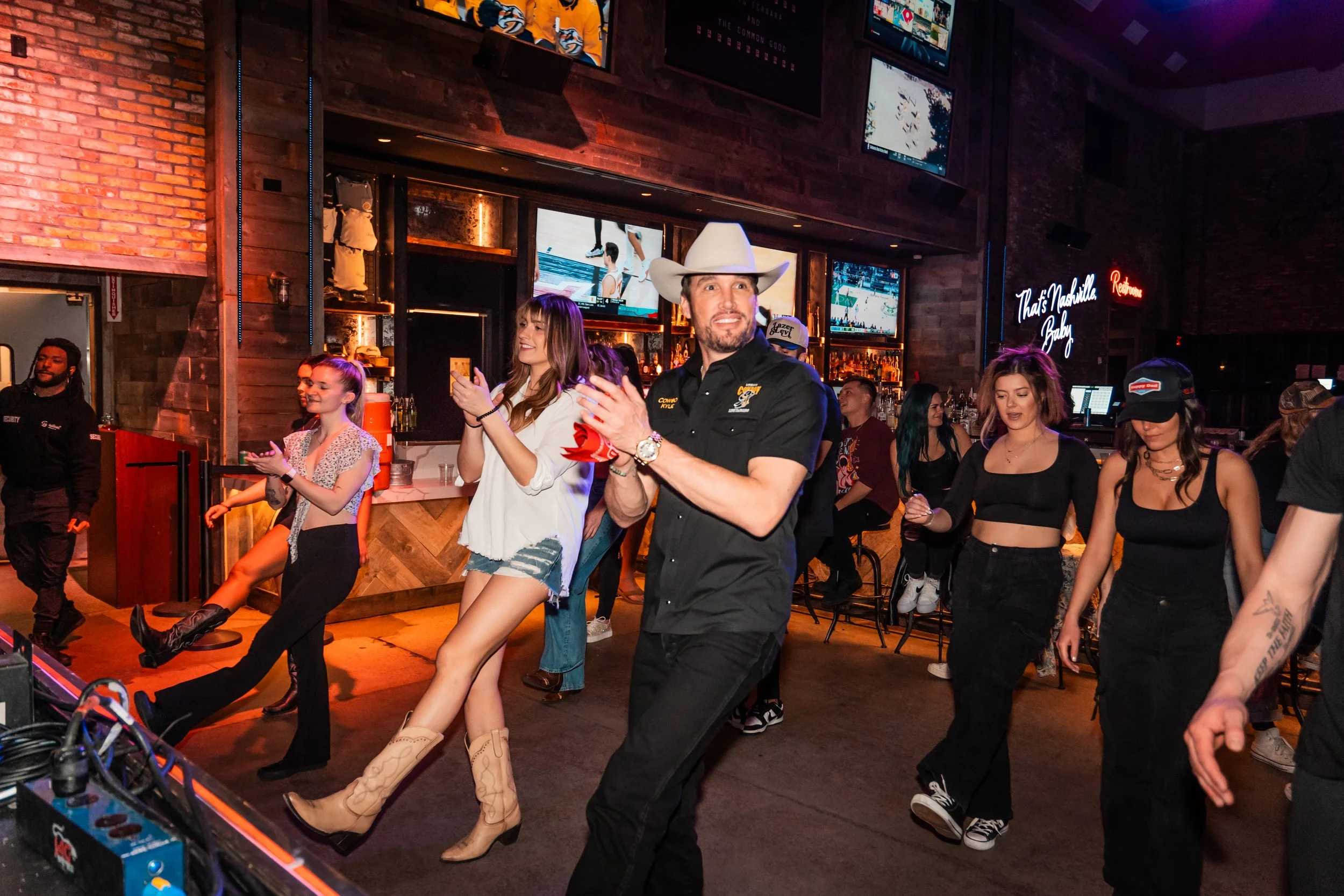 People dancing and socializing in a dimly lit bar with multiple TV screens, a neon sign reading 'Texas Mashkill Baby,' and a brick wall background.