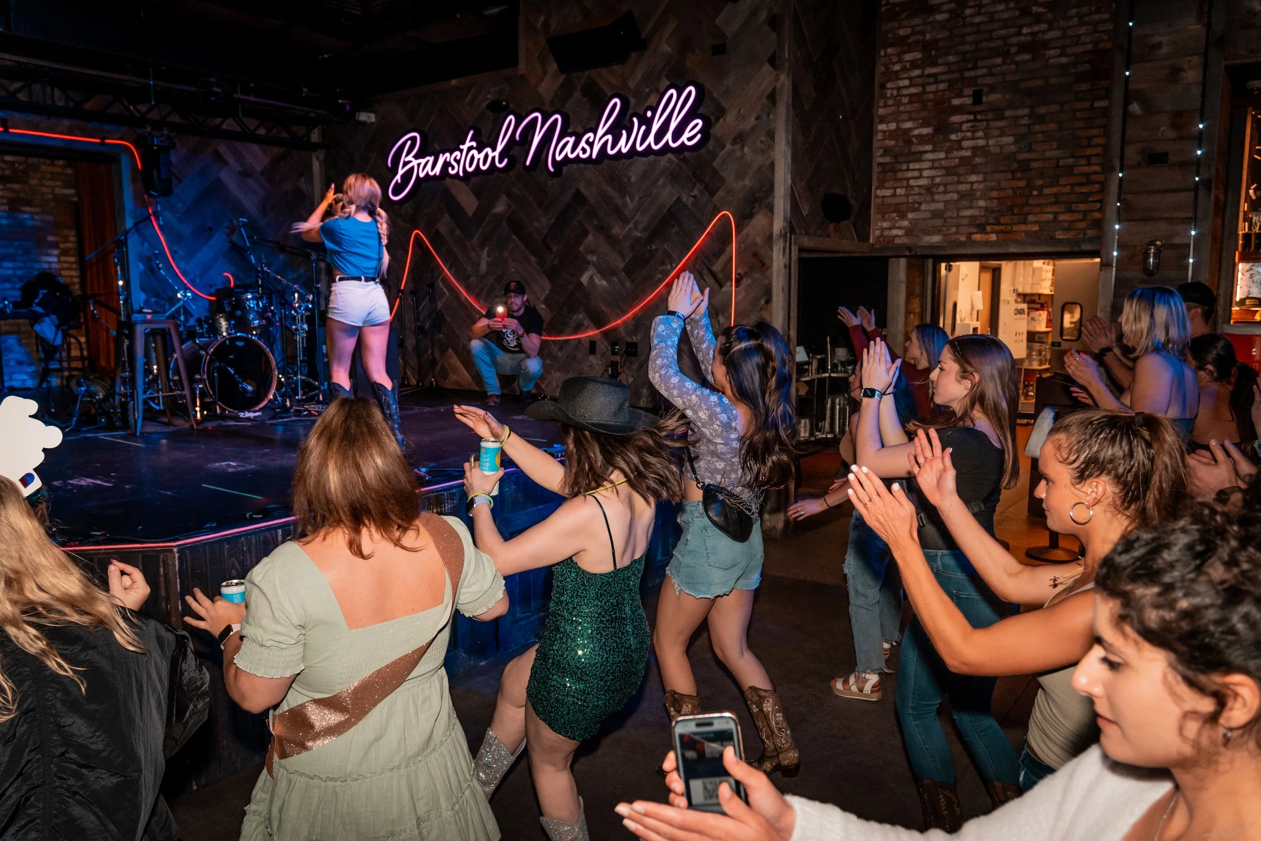 Dance floor at Barstool Nashville with a performer on stage, audience members clapping and enjoying the show, neon sign reading 'Barstool Nashville' on brick wall, and people taking photos.