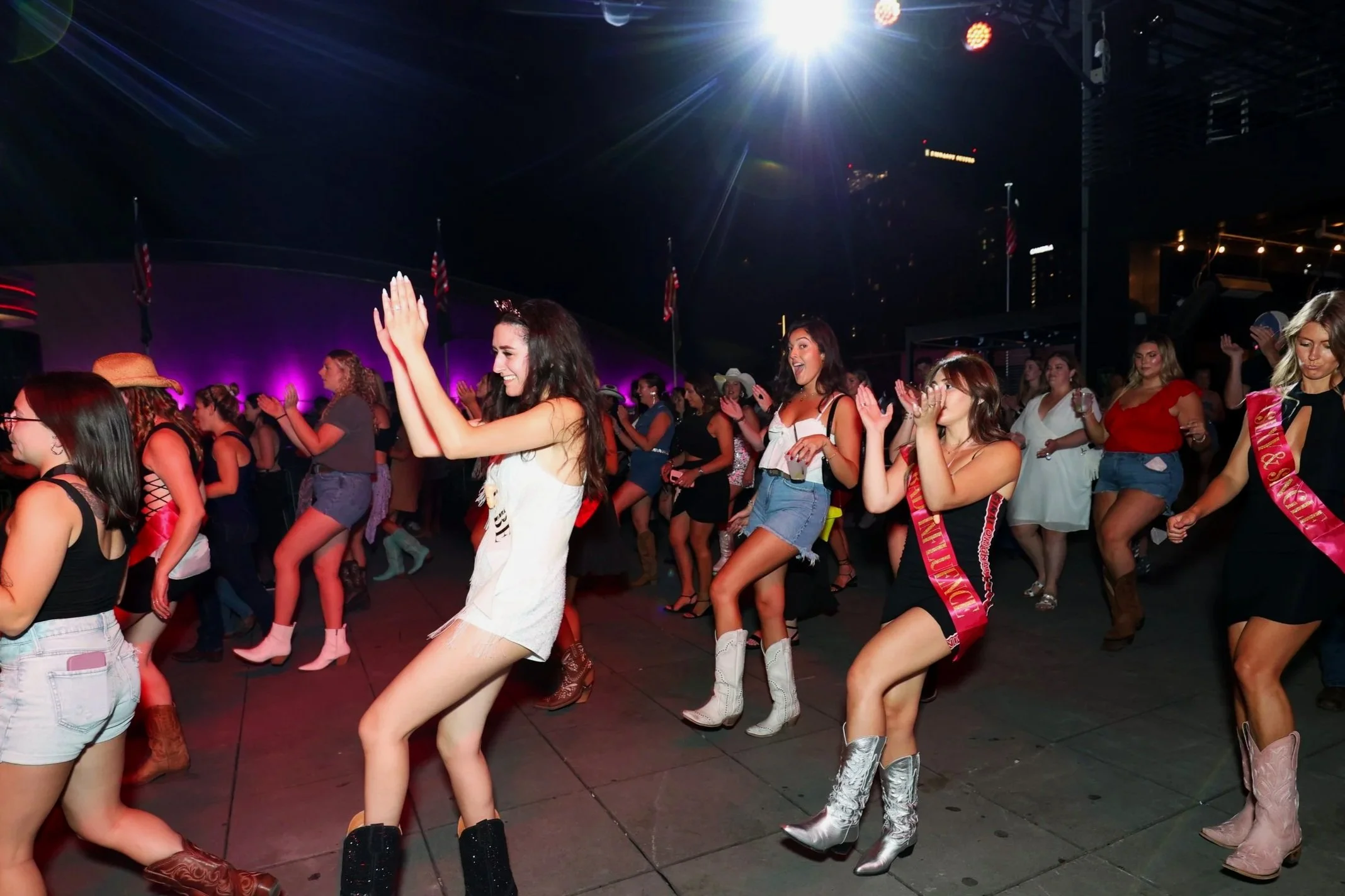 A group of women dancing at a lively indoor event with purple and pink lighting, some wearing sashes and cowboy boots.