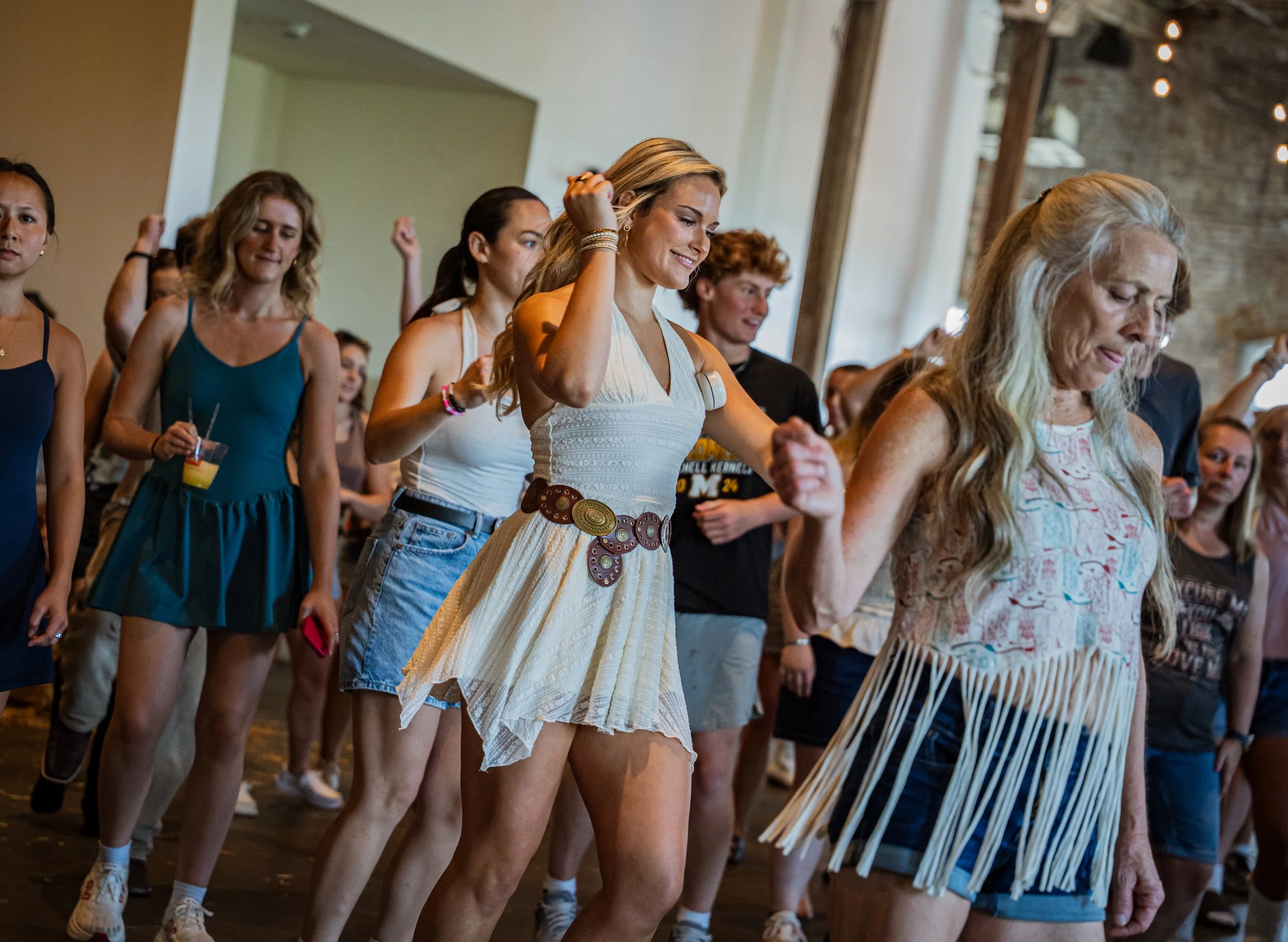 A group of young women dancing at an indoor event, holding hands and smiling, in a spacious venue with wooden beams and string lights.