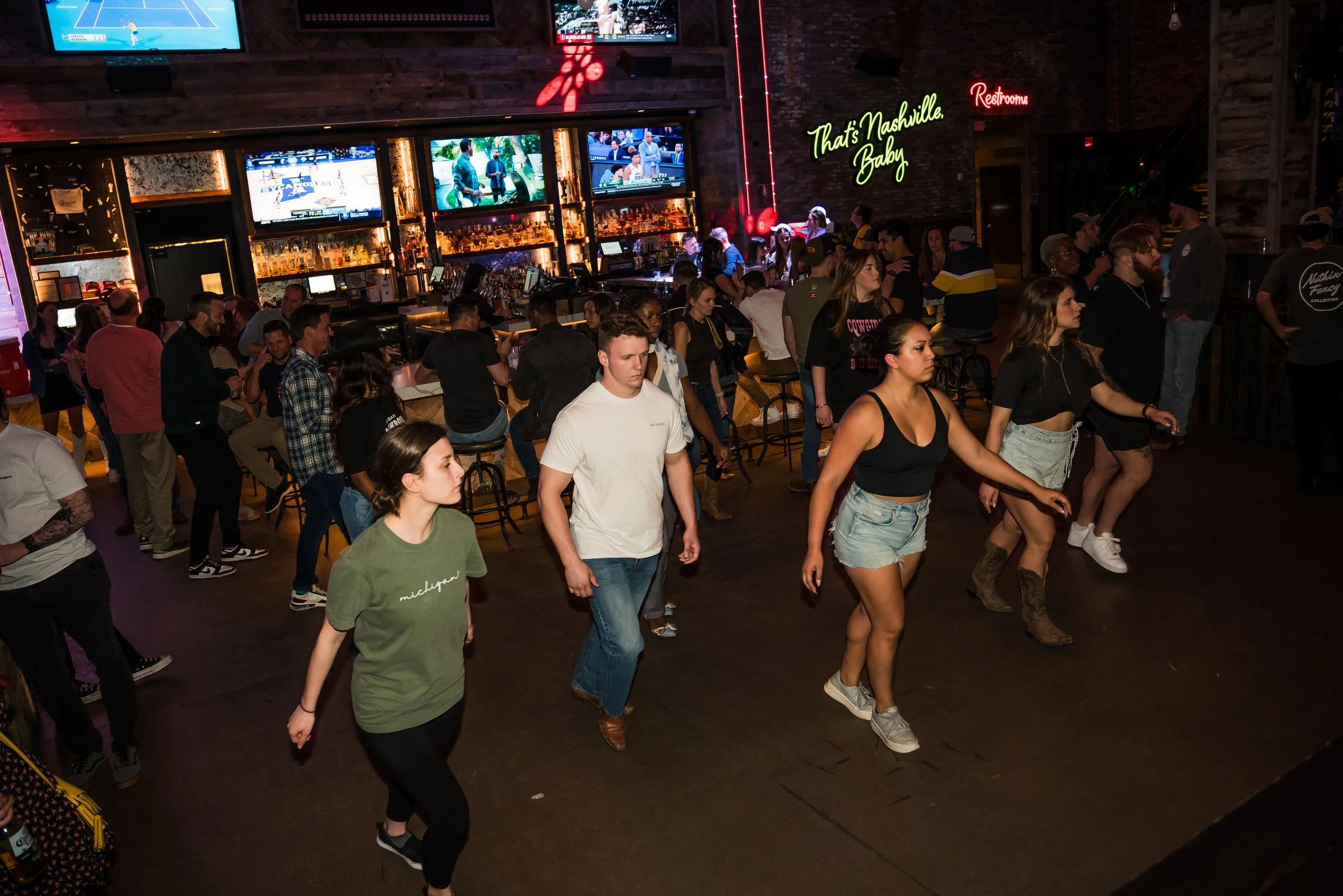 A crowded indoor bar or nightclub with people dancing, socializing, and watching sports on large screens. Neon signs display 'That's Nashville Baby' and 'Restrooms'.