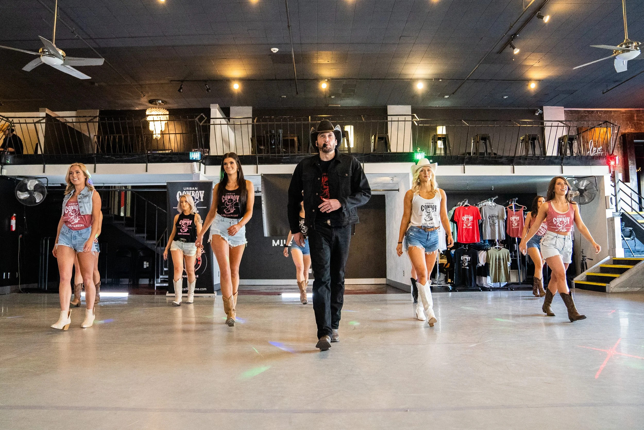 Group of women and man walking in a line in a dance studio, with a banner reading 'Urban Cowboy' behind them.