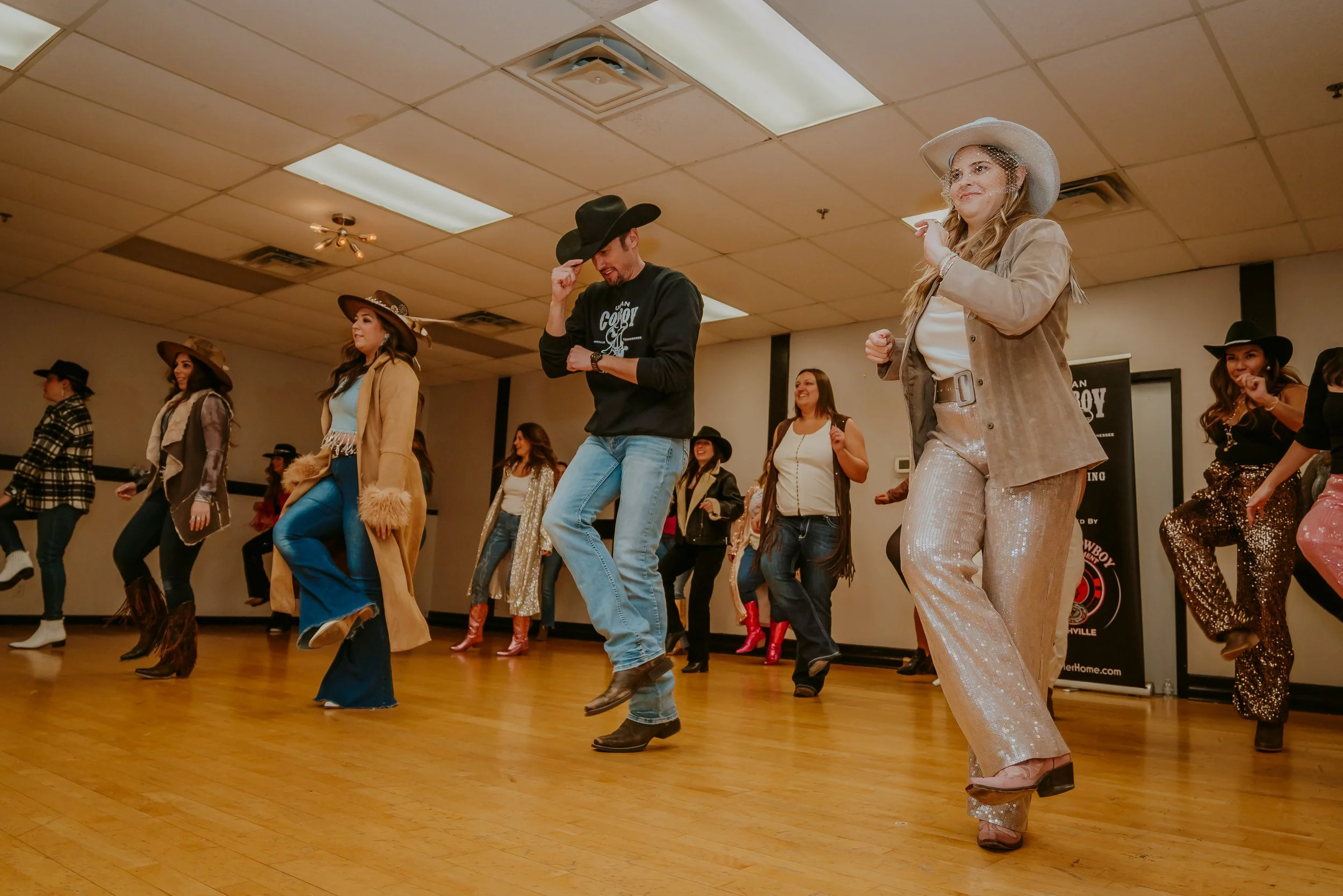 Group of people participating in a line dance wearing country western attire, including cowboy hats, boots, and sequined outfits, in a dance hall with wood floors and a ceiling with fluorescent lights.