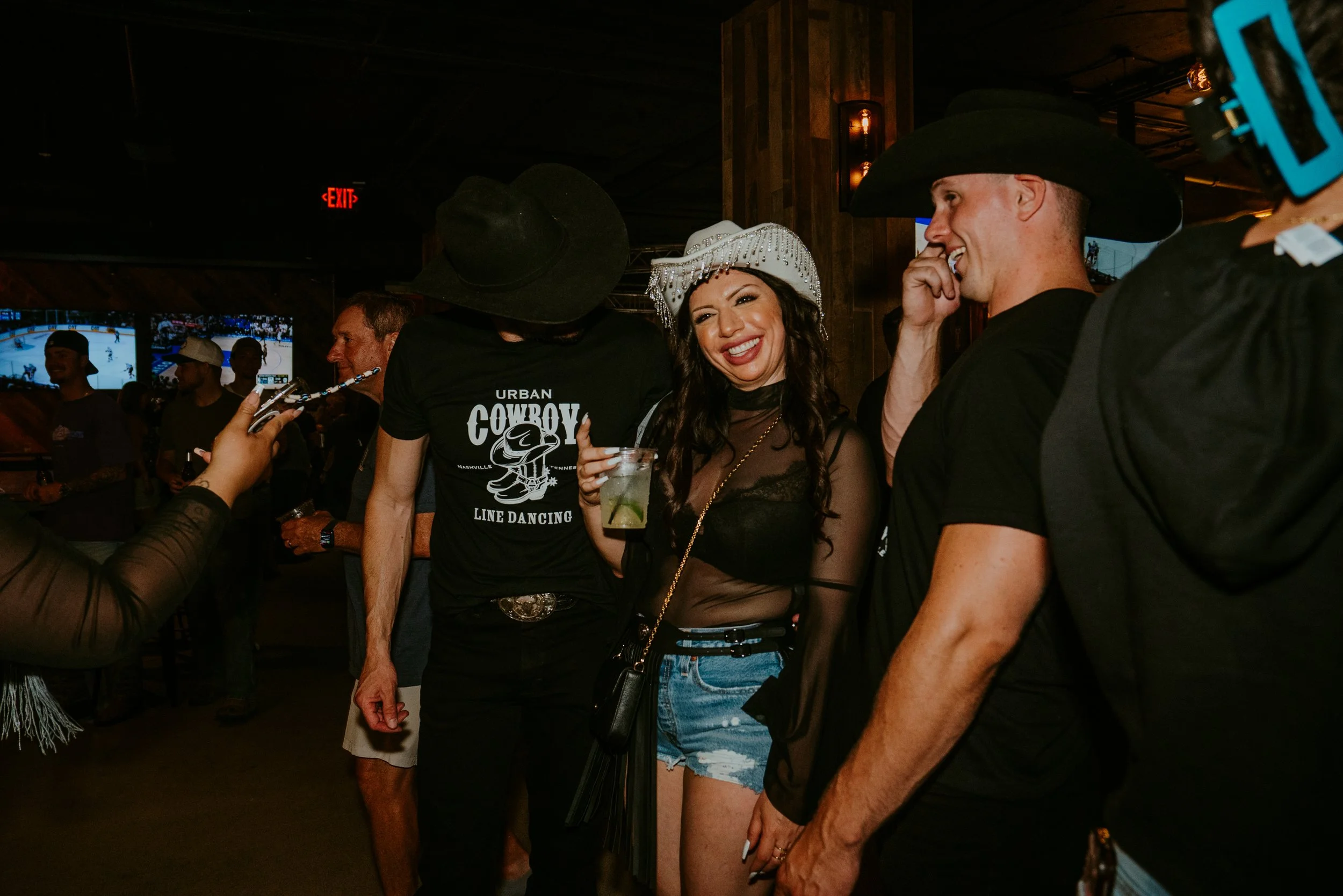 A group of six people in cowboy hats socializing in a dimly lit bar or restaurant, with multiple television screens showing ice hockey in the background.