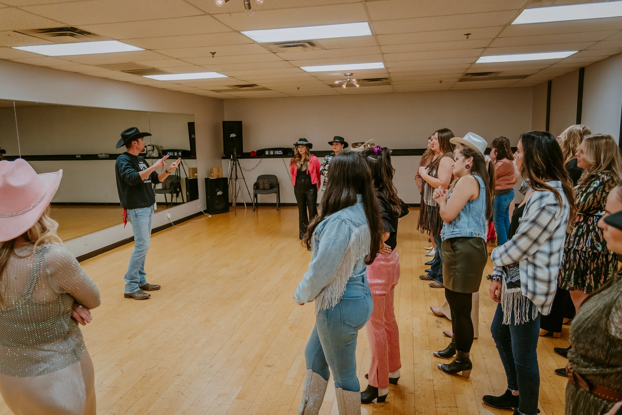 A group of diverse women dressed in western and cowgirl costumes, standing in a dance studio, participating in a line dance class led by an instructor wearing a cowboy hat and casual clothing.