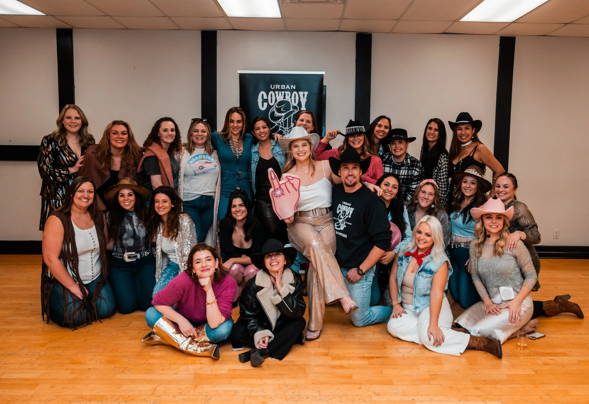A group of women and a man dressed in western and cowgirl-themed outfits, posing for a group photo on a wooden floor in front of a plain wall with a banner that reads "URBAN COWBOY" and features a cowboy hat logo.
