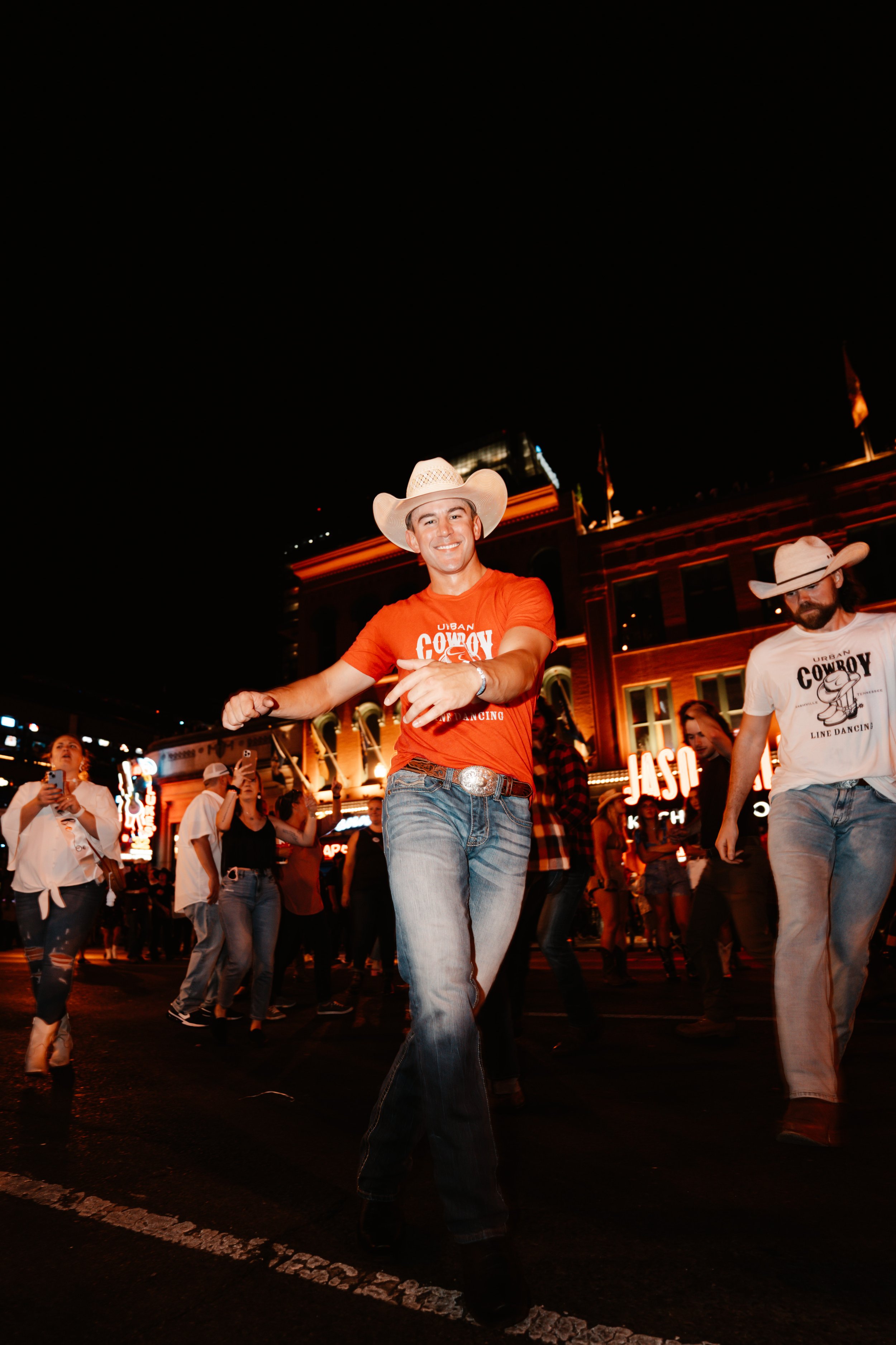 A group of people dancing outside at night, with two men wearing cowboy hats in the foreground, one smiling and pointing, the other looking down. The scene is illuminated by artificial lights with a building in the background.