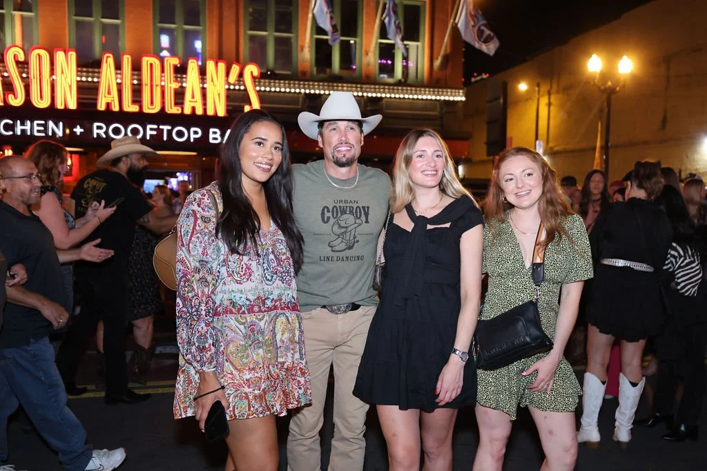 Four people posing in front of a restaurant with a neon sign at night, with a crowd in the background.