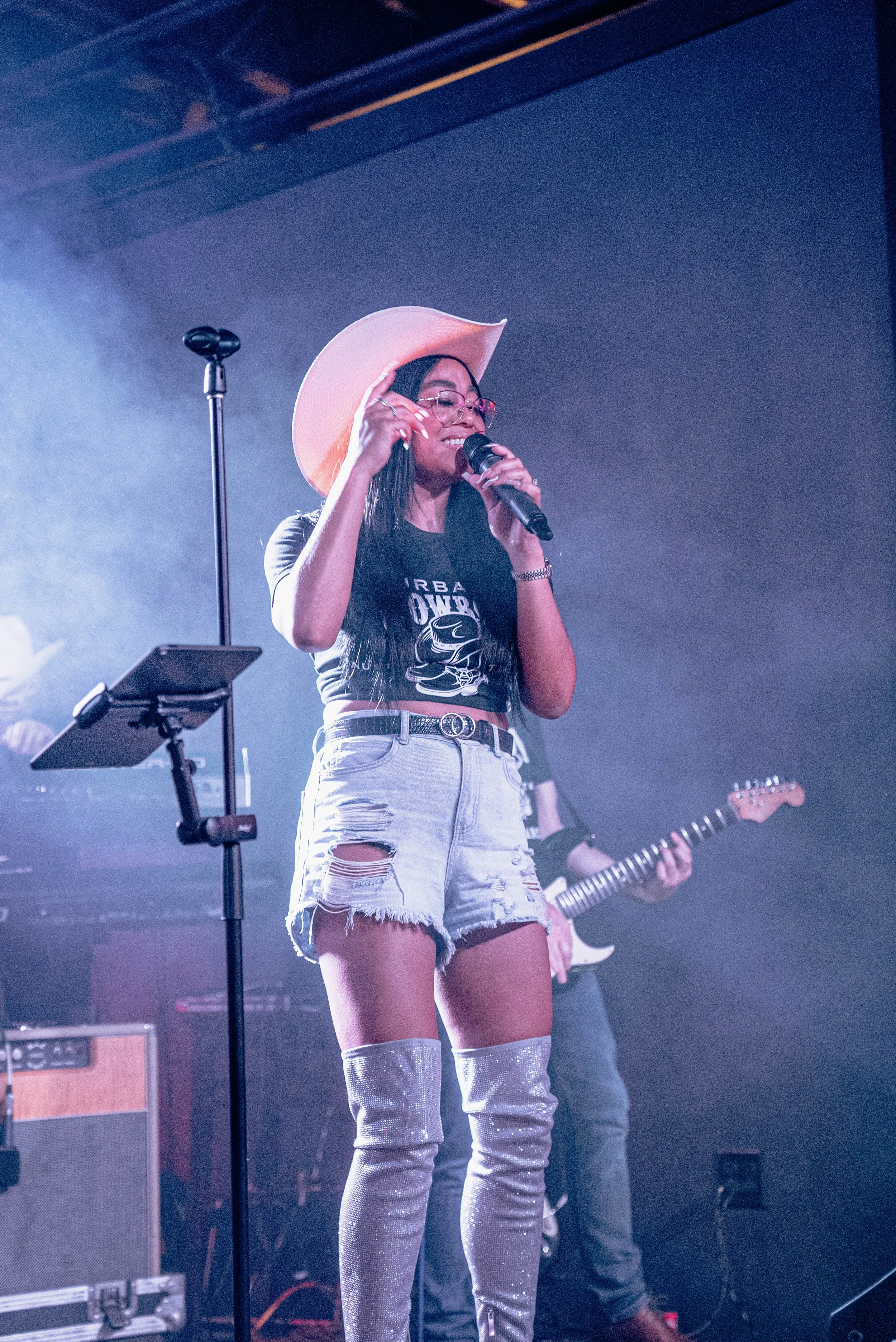 A woman wearing a large pink hat, glasses, a black t-shirt with the text 'URBAN' and high-waisted, ripped denim shorts, singing into a microphone on stage with a guitarist in the background and music equipment around.
