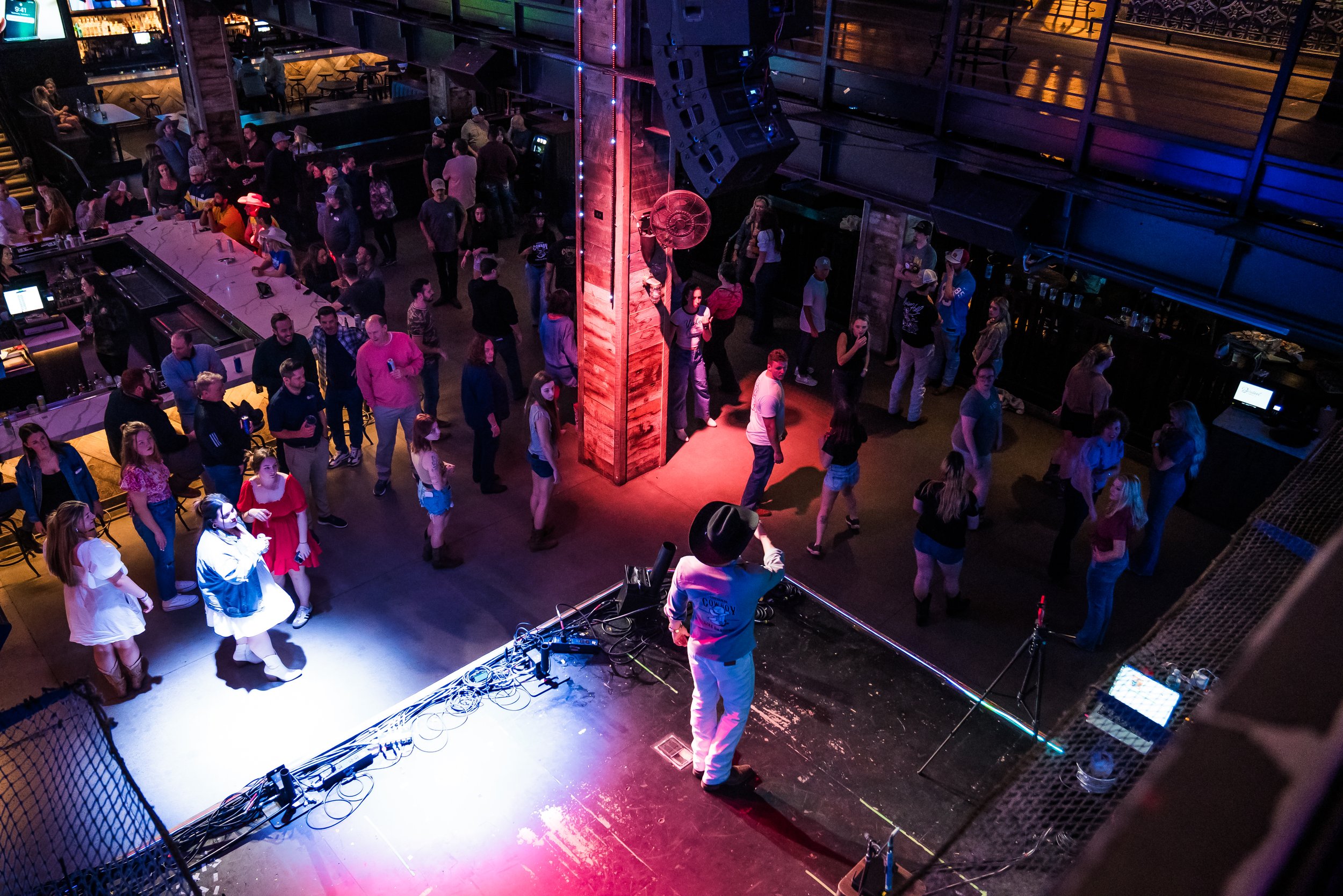 The image shows an indoor event with a dance floor filled with people. There is a DJ booth with equipment at the front, and a stage with large speakers and colorful lighting. Some people are dancing, while others are standing or sitting around. The v