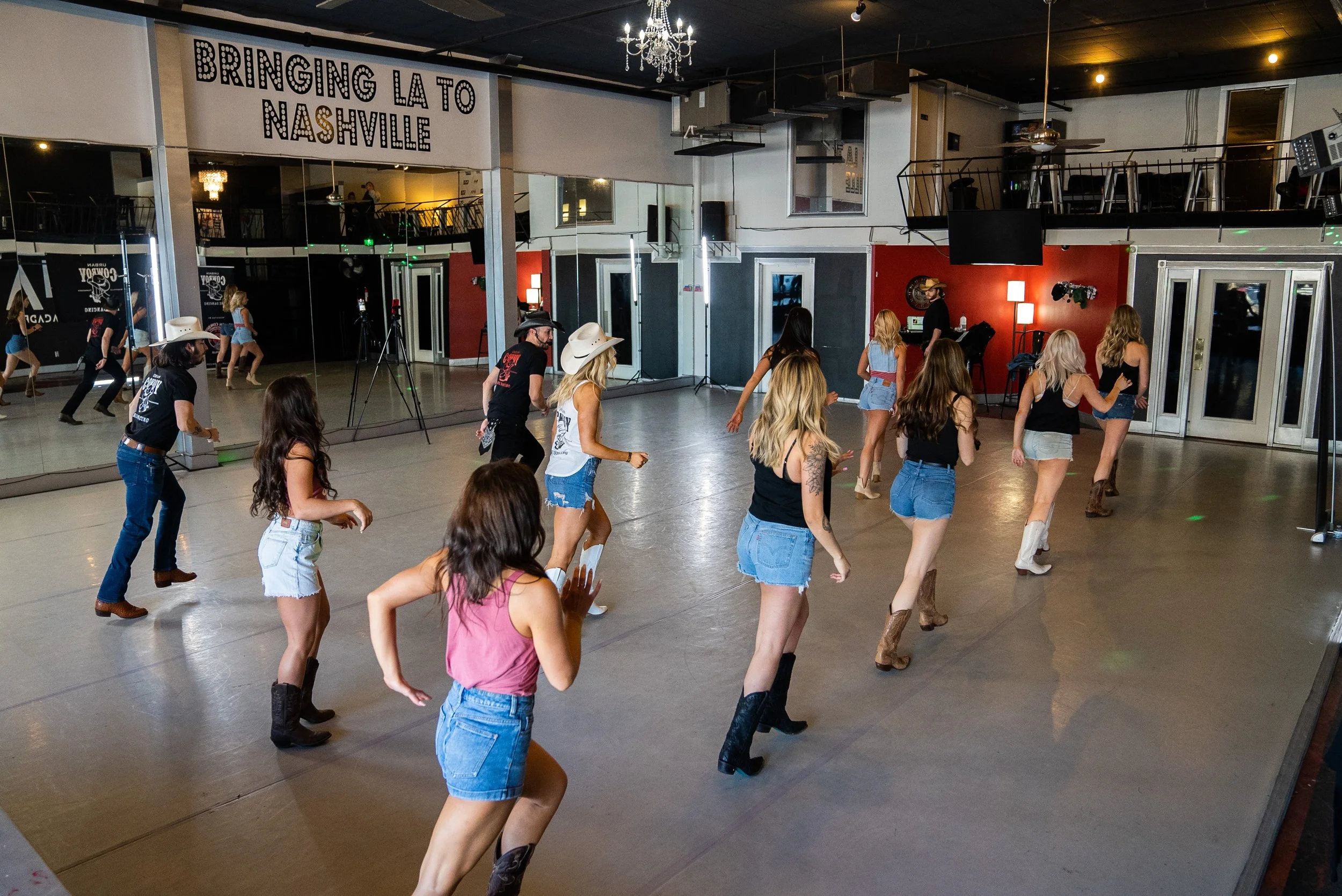 People line dancing in a spacious indoor dance studio with a mirror wall, chandelier, and a sign reading 'Bringing LA to Nashville'