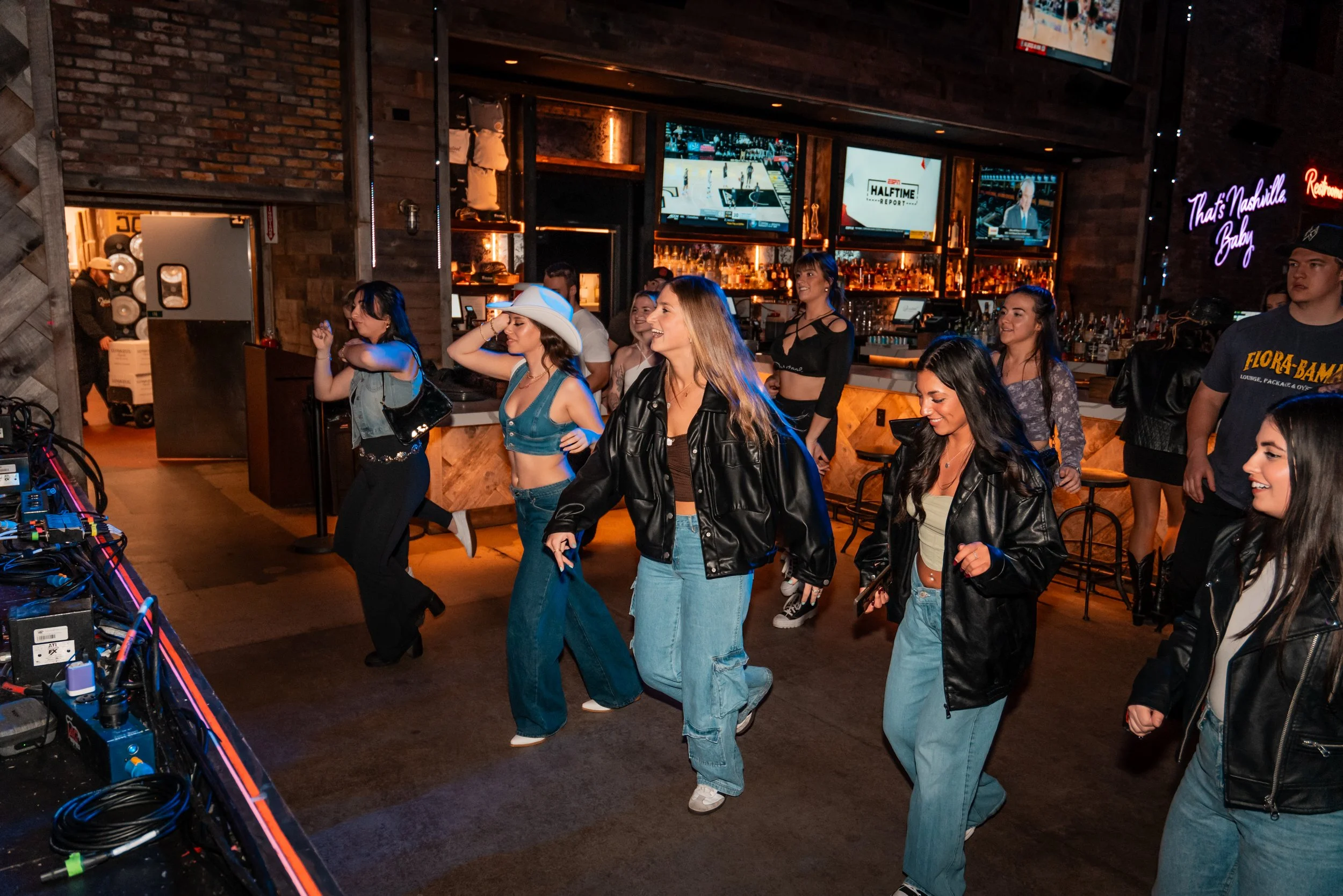 Group of young women dancing and having fun at a bar with multiple television screens and a neon sign that reads "That's Nashville Baby".