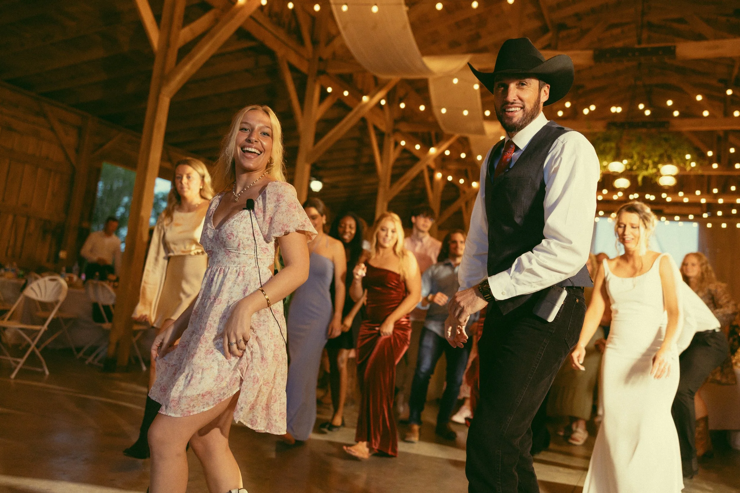 People dancing at a wedding or party inside a wooden venue decorated with string lights.
