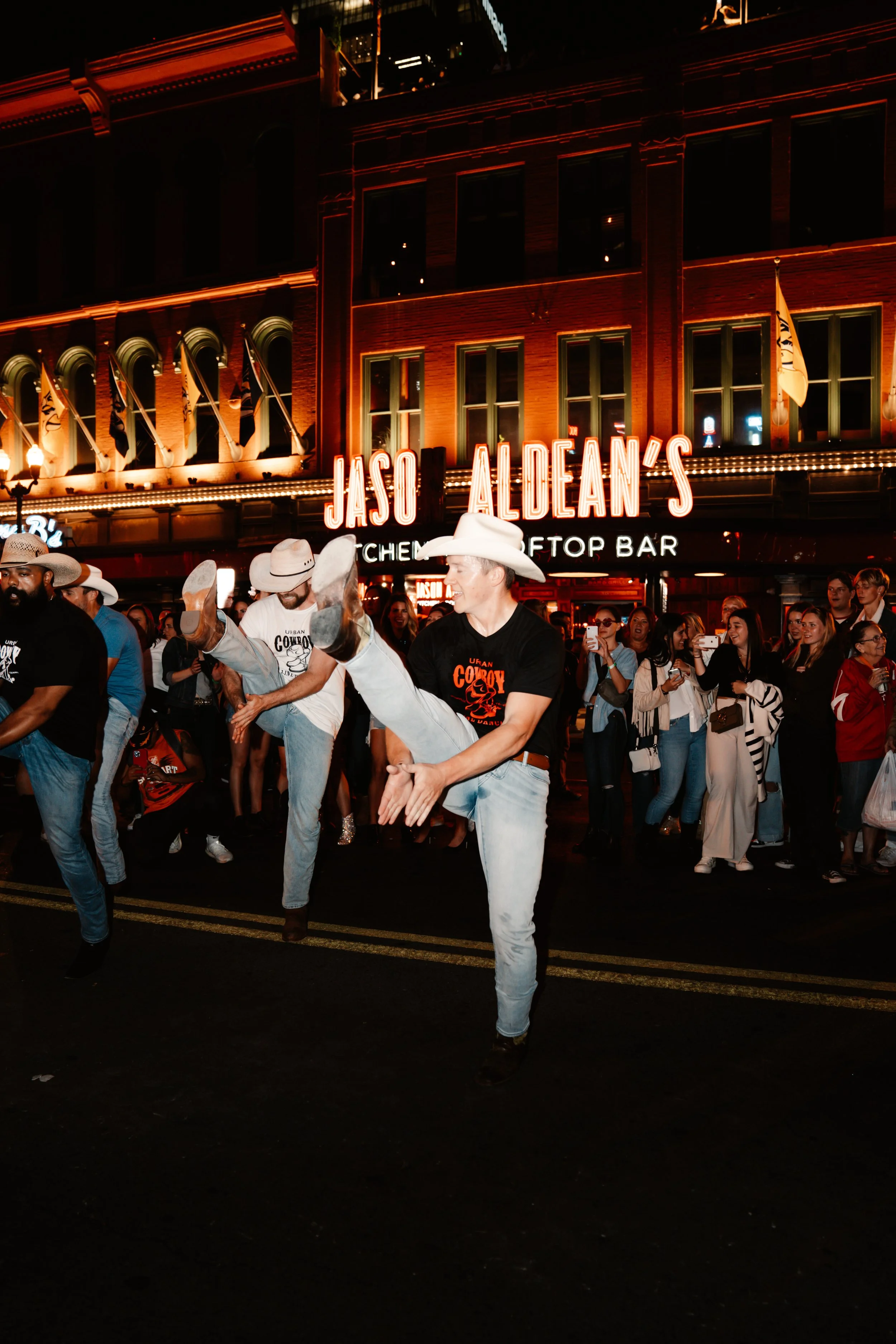 A lively street scene at night in front of Jaso Aldean's restaurant and rooftop bar, with people dancing and enjoying themselves, some wearing cowboy hats.