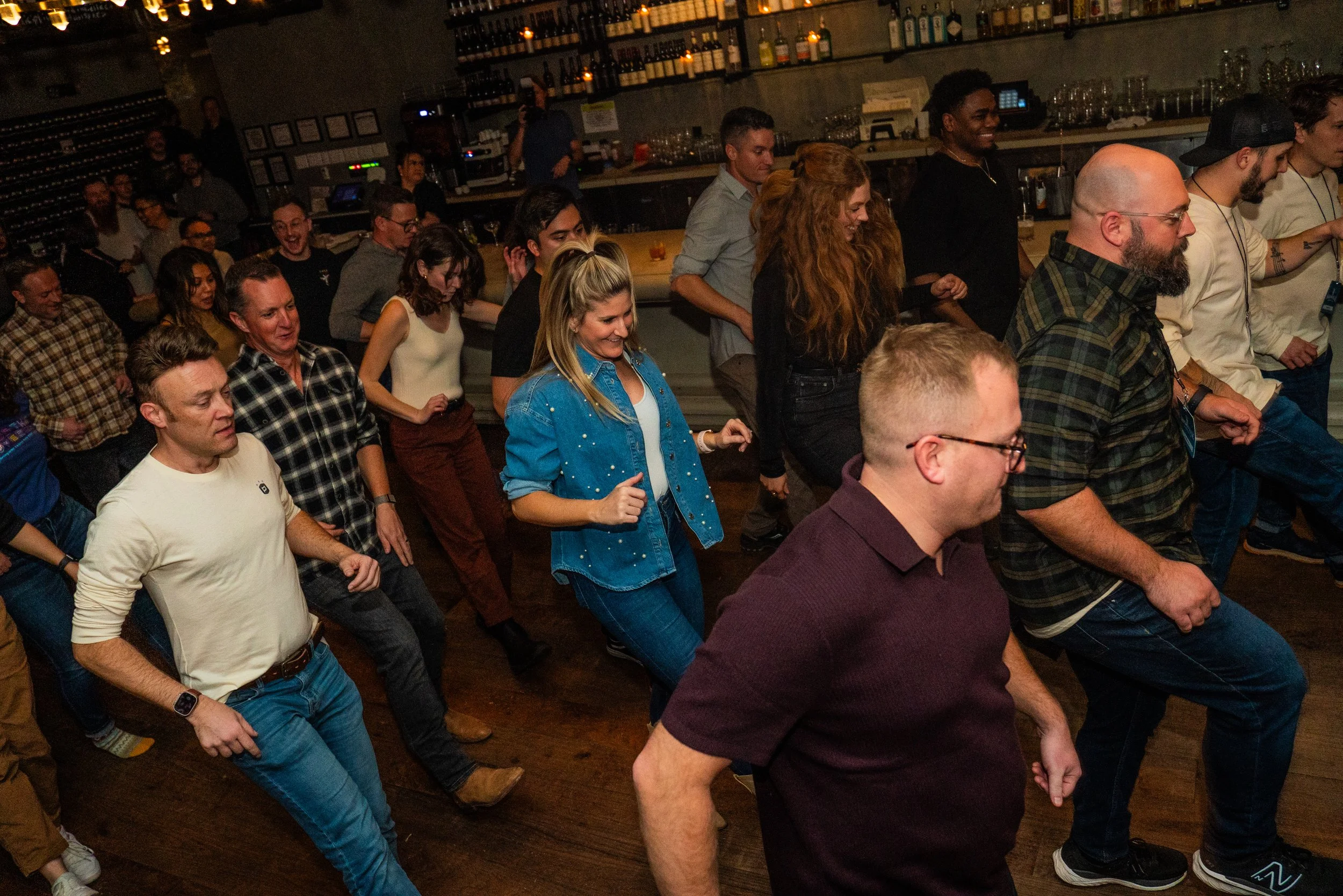 People dancing at a bar or club, with a bar and shelves of liquor bottles in the background.