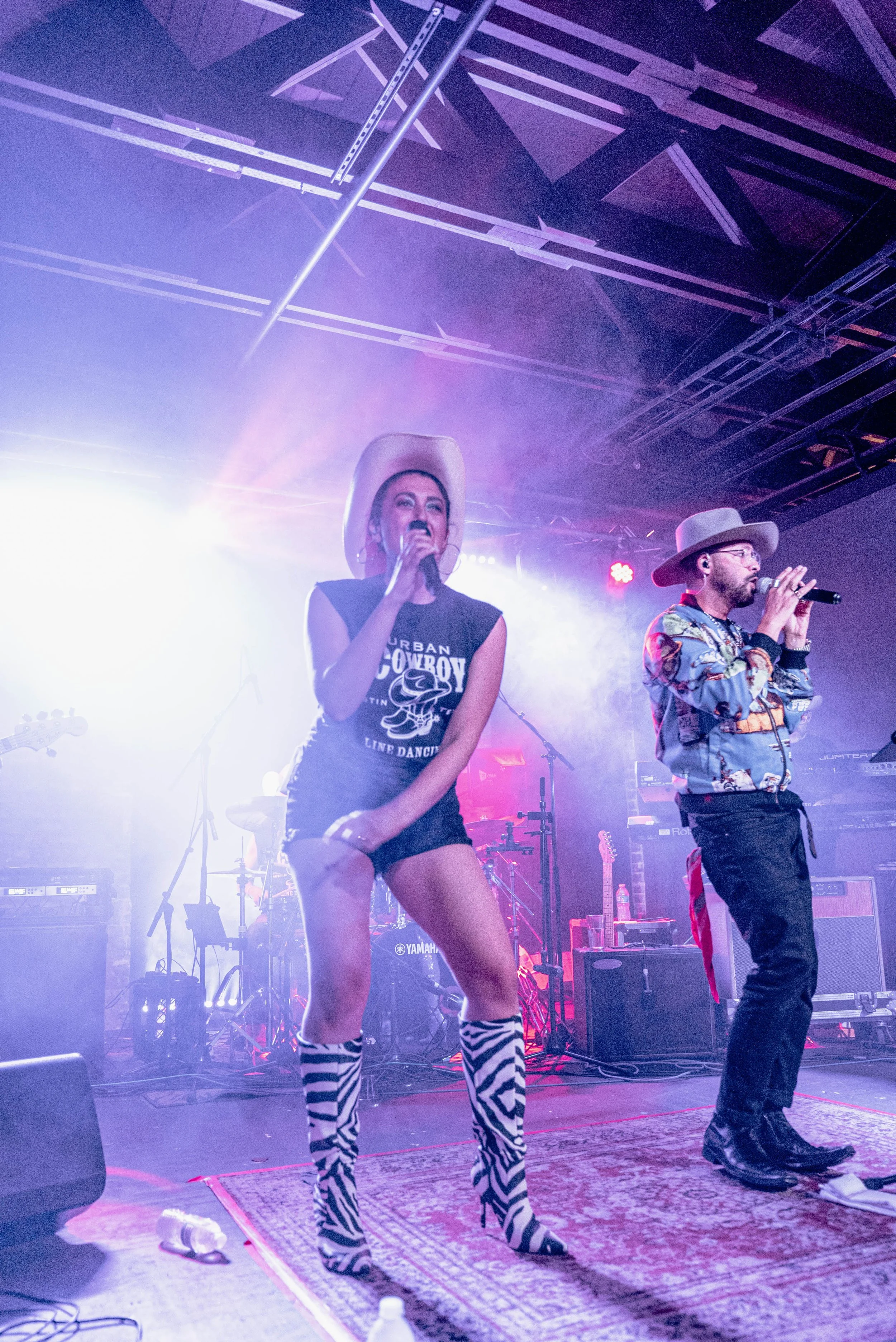 Two singers perform on stage with musical instruments and equipment in the background, under vibrant stage lighting and smoke effects, in an indoor concert setting.
