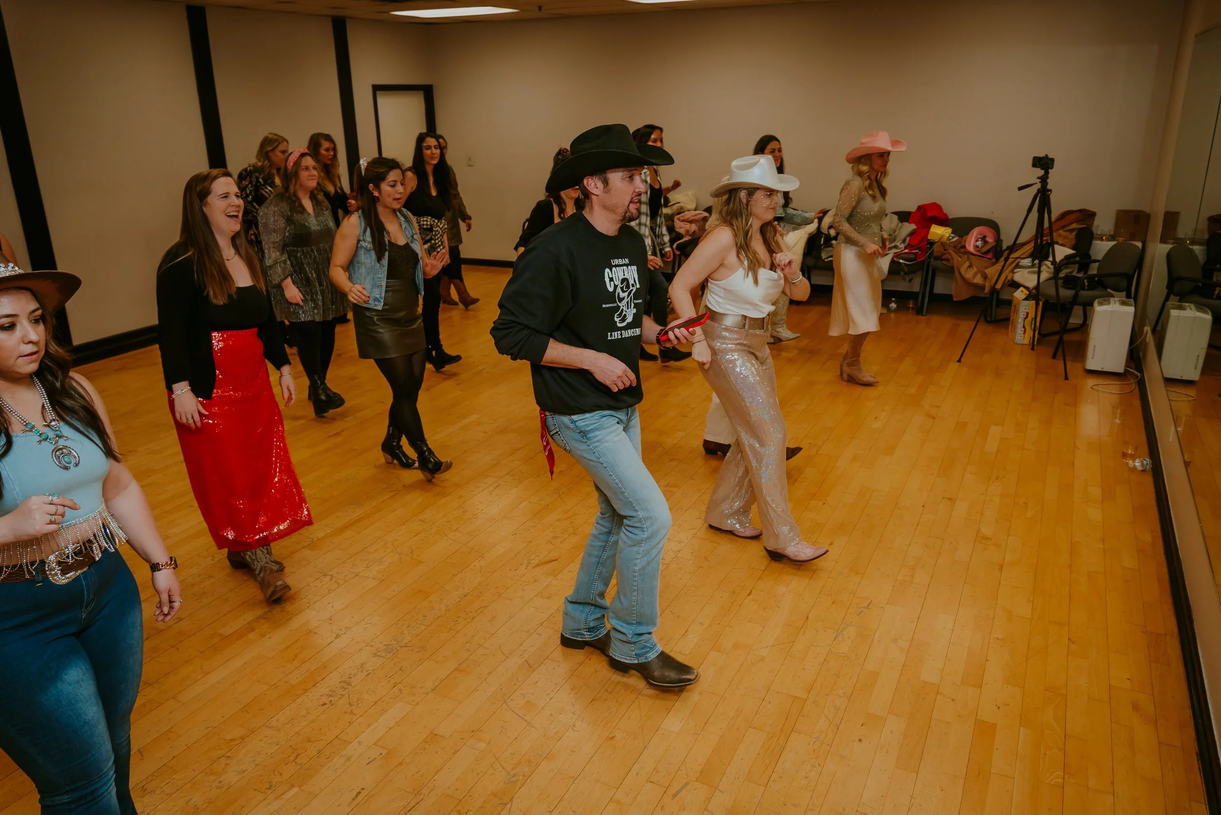 A group of women and two men line dancing in a room with wooden floors, some wearing cowboy hats, western-style clothing, and boots. There is a camera on a tripod and chairs against the wall with miscellaneous items on them.