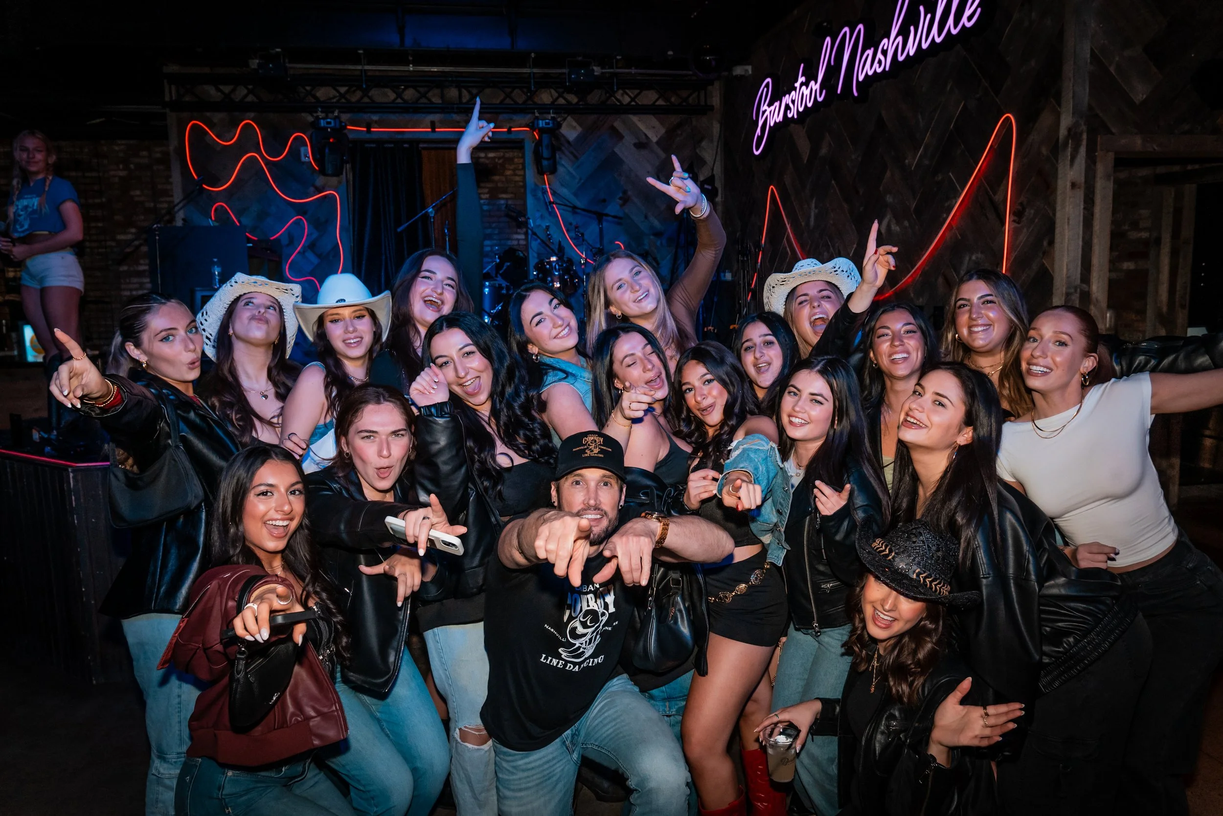 Group of young women and one man posing for a photo at a lively bar or nightclub. Some women are wearing cowboy hats. The background features neon lights and a sign that says 'Barstool Nashville.' Everyone appears to be having fun.
