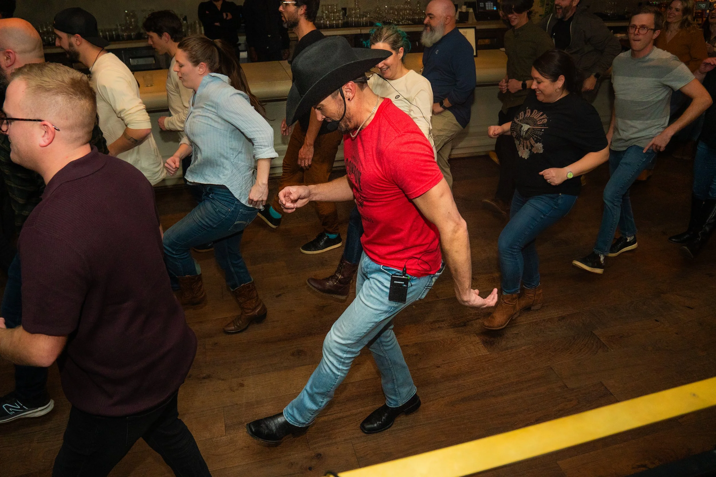A group of people dancing in a line at an indoor social gathering or party. Some are wearing cowboy boots and hats, and they appear to be enjoying themselves.