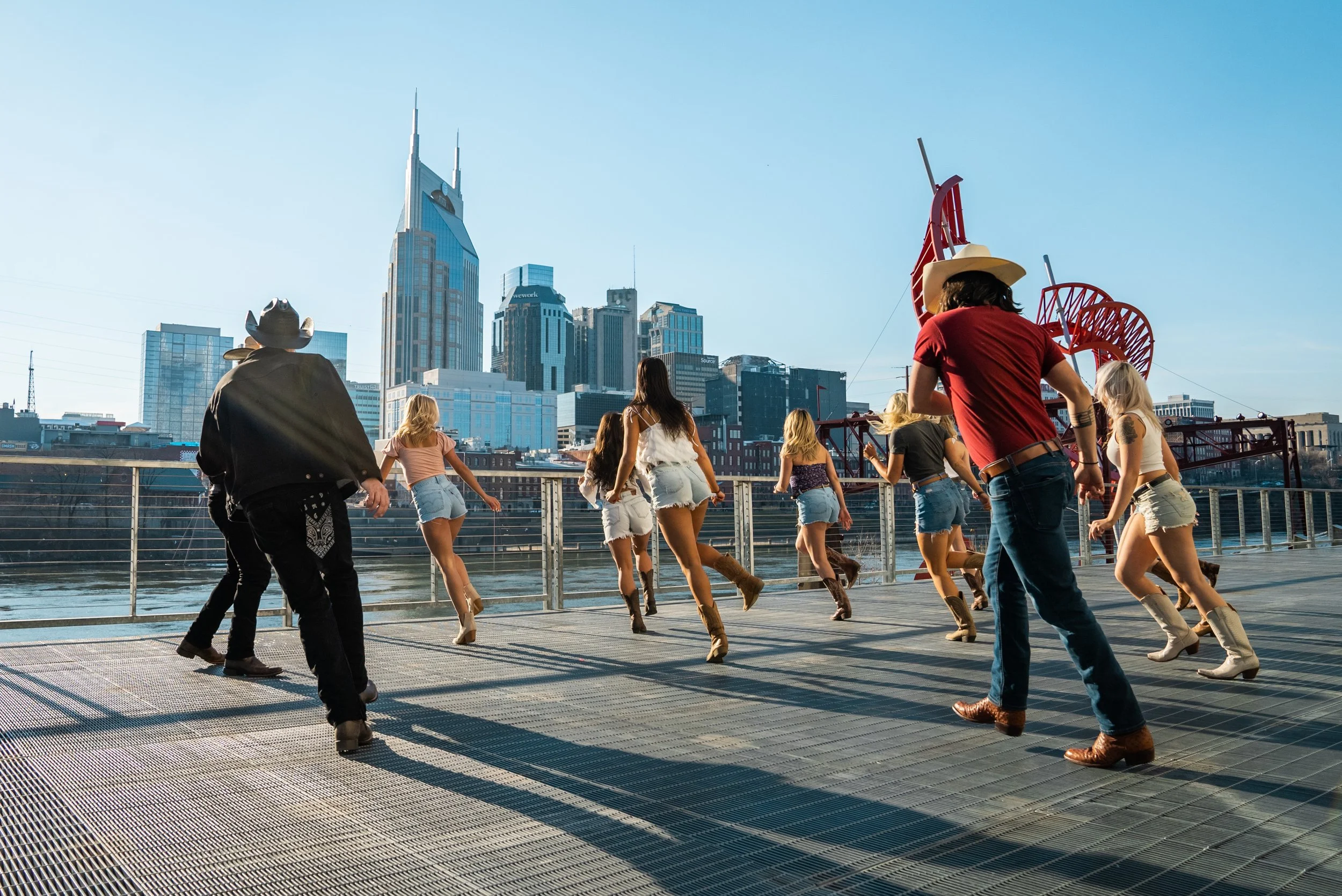 People line dance on a rooftop with a city skyline in the background, including the Aerial tramway, with tall buildings and a clear blue sky.
