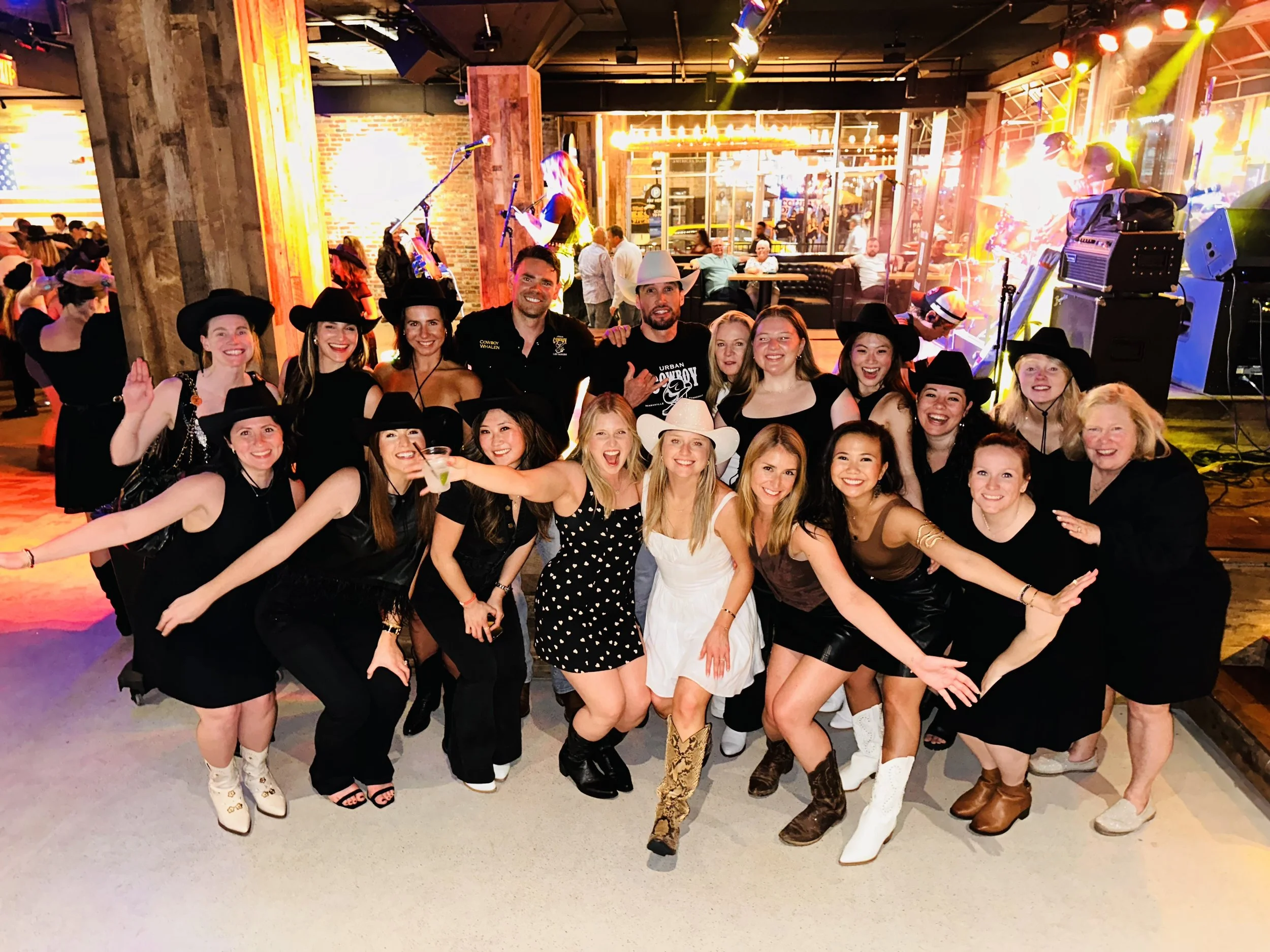 A large group of people, mostly women, dressed in cowboy and cowgirl outfits, posing together at a lively indoor event or party, with a stage and band performing in the background, colorful lights, and a brick wall behind them.