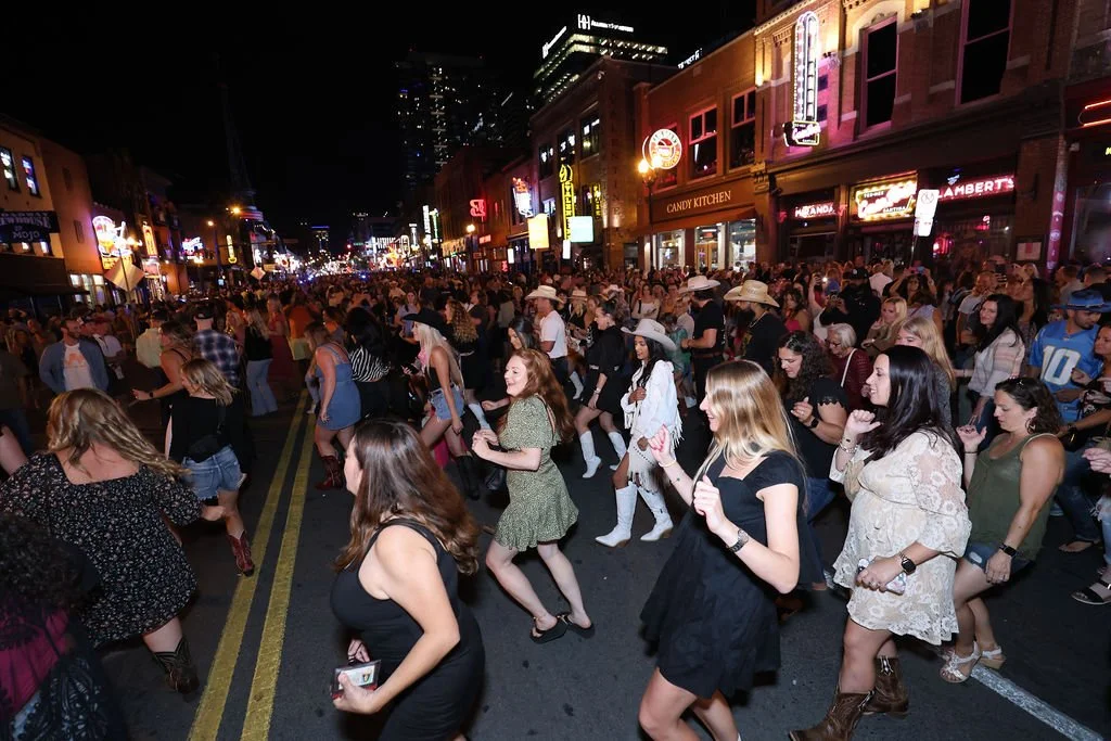 People dancing in the street at night during a lively outdoor event, with neon signs and storefronts in the background.
