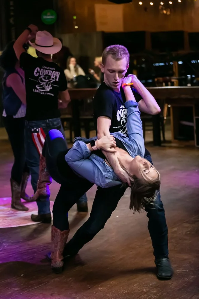 A man and a woman are dancing in a dimly lit bar or club. The man is holding the woman in a dip, and she is leaning back with her eyes closed. Both are wearing black shirts with 