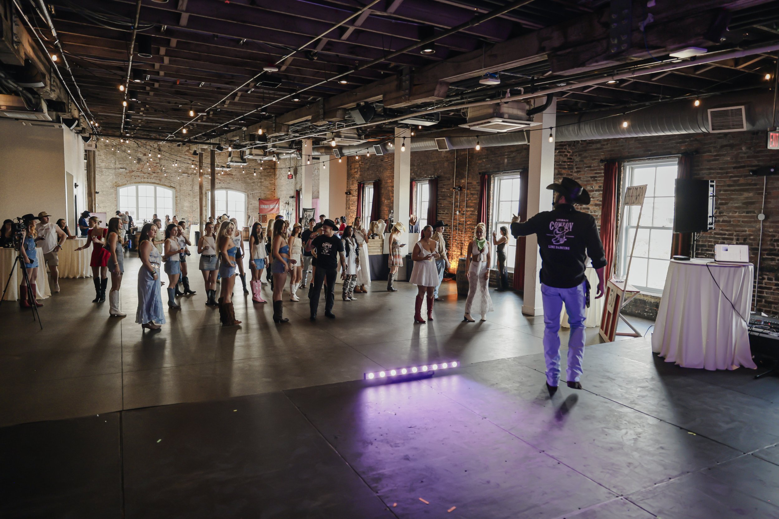 Interior of a rustic-style event space with exposed brick and wooden beams, where a line dance class is taking place, and a woman in cowboy attire is leading the group.