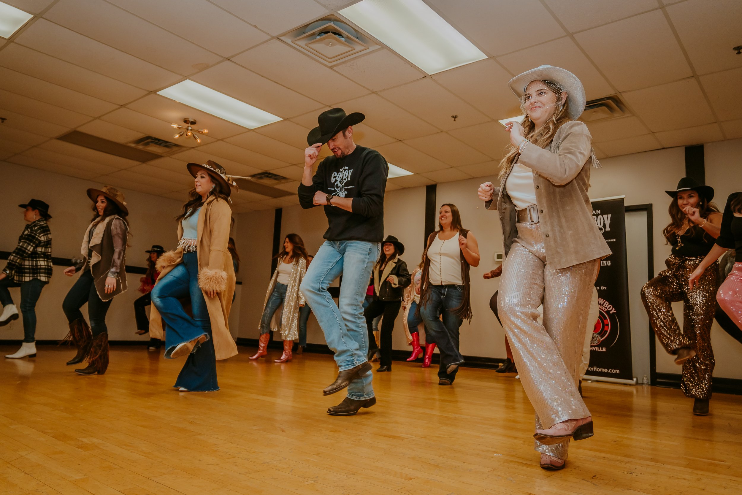 Group of people dressed in western and country-themed outfits, dancing in a line in a room with wooden floors and ceiling lights.