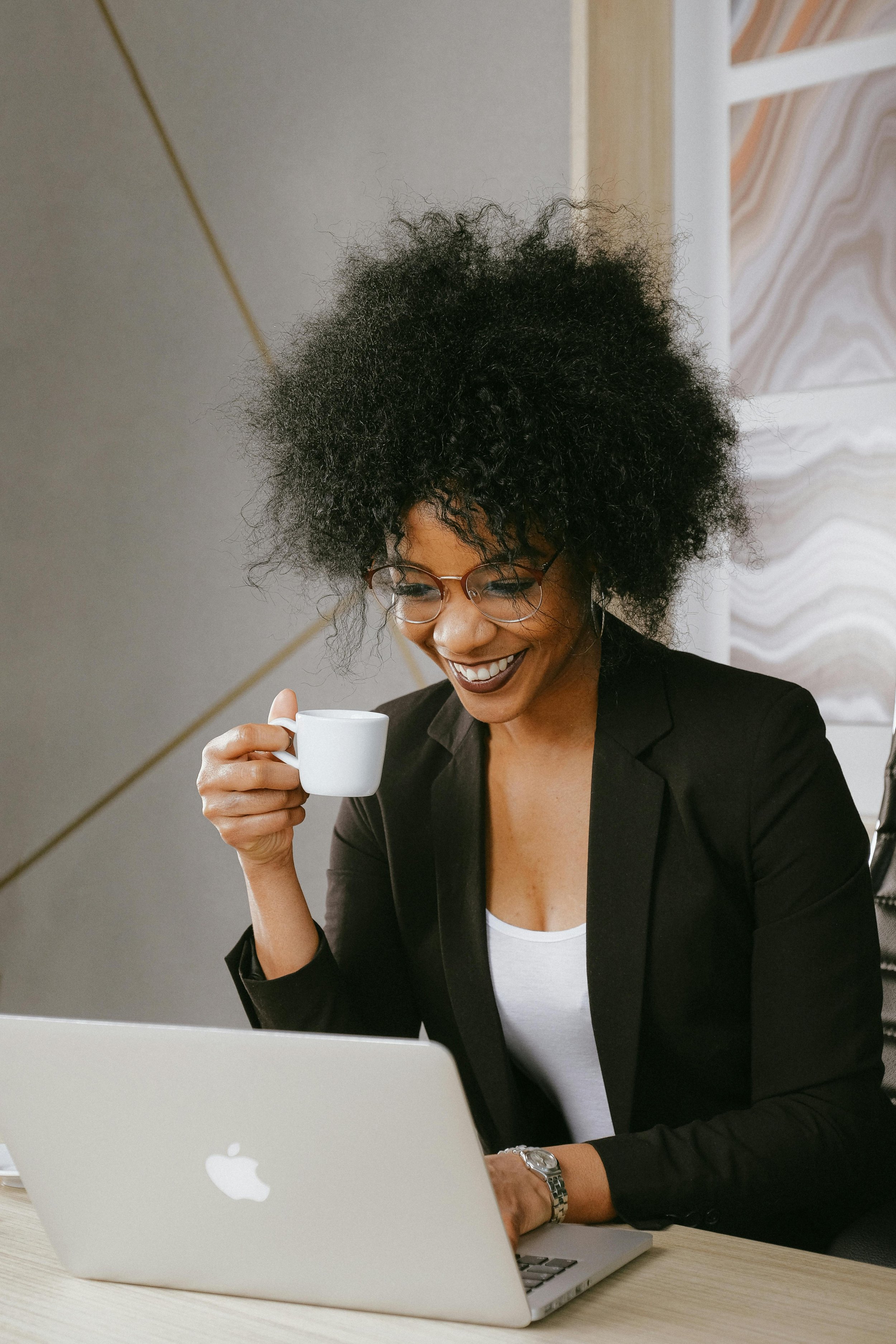 Smiling woman wearing a black jacket and white top, holding a white coffee cup and smiling while looking at laptop.