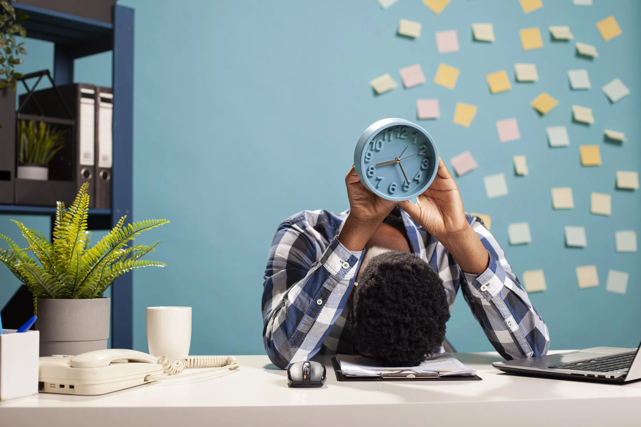 Man in plaid shirt with his head on a desk, holding a clock over his head. The blue wall behind him is covered in post it notes