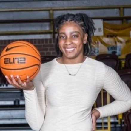 A woman standing in a sports arena holding a basketball and smiling at the camera.