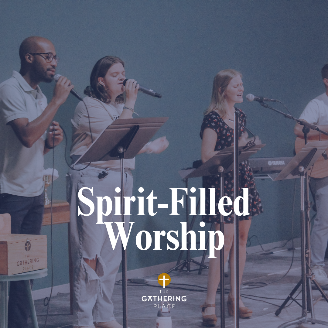 A group of four people performing music on stage, with an audience watching them. The performers include a man with a microphone and a keyboard, a man with glasses standing and smiling, a man with a guitar, and a woman singing into a microphone, all in an indoor setting.