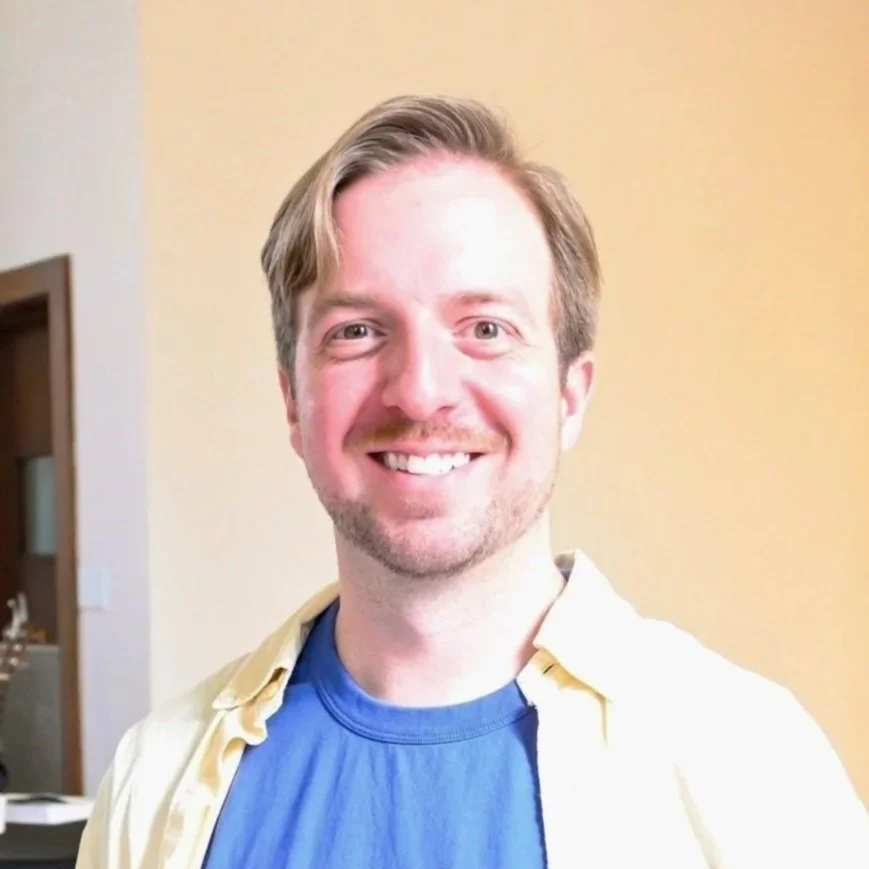 A smiling man with light brown hair, wearing a blue shirt and light yellow jacket, standing inside a room with a beige wall in the background.