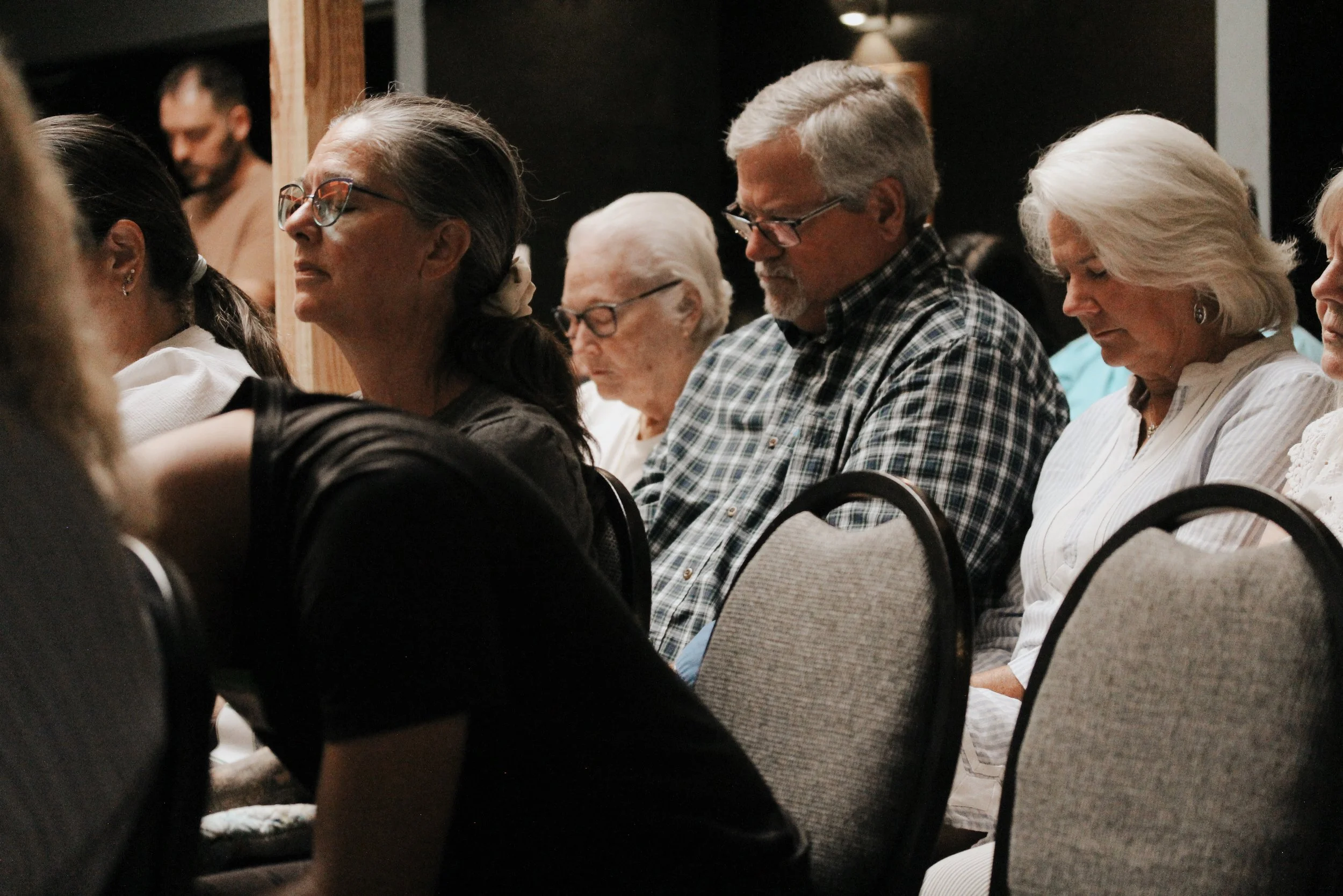 People sitting in a row at an indoor event, with some appearing to be praying or meditating, mostly with eyes closed and heads bowed.