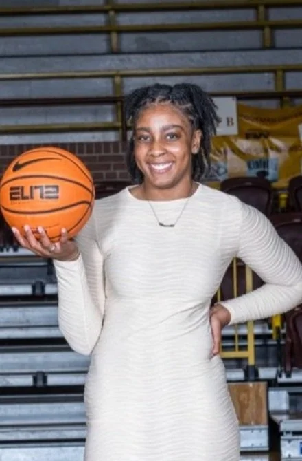 A woman standing in a sports arena holding a basketball and smiling at the camera.
