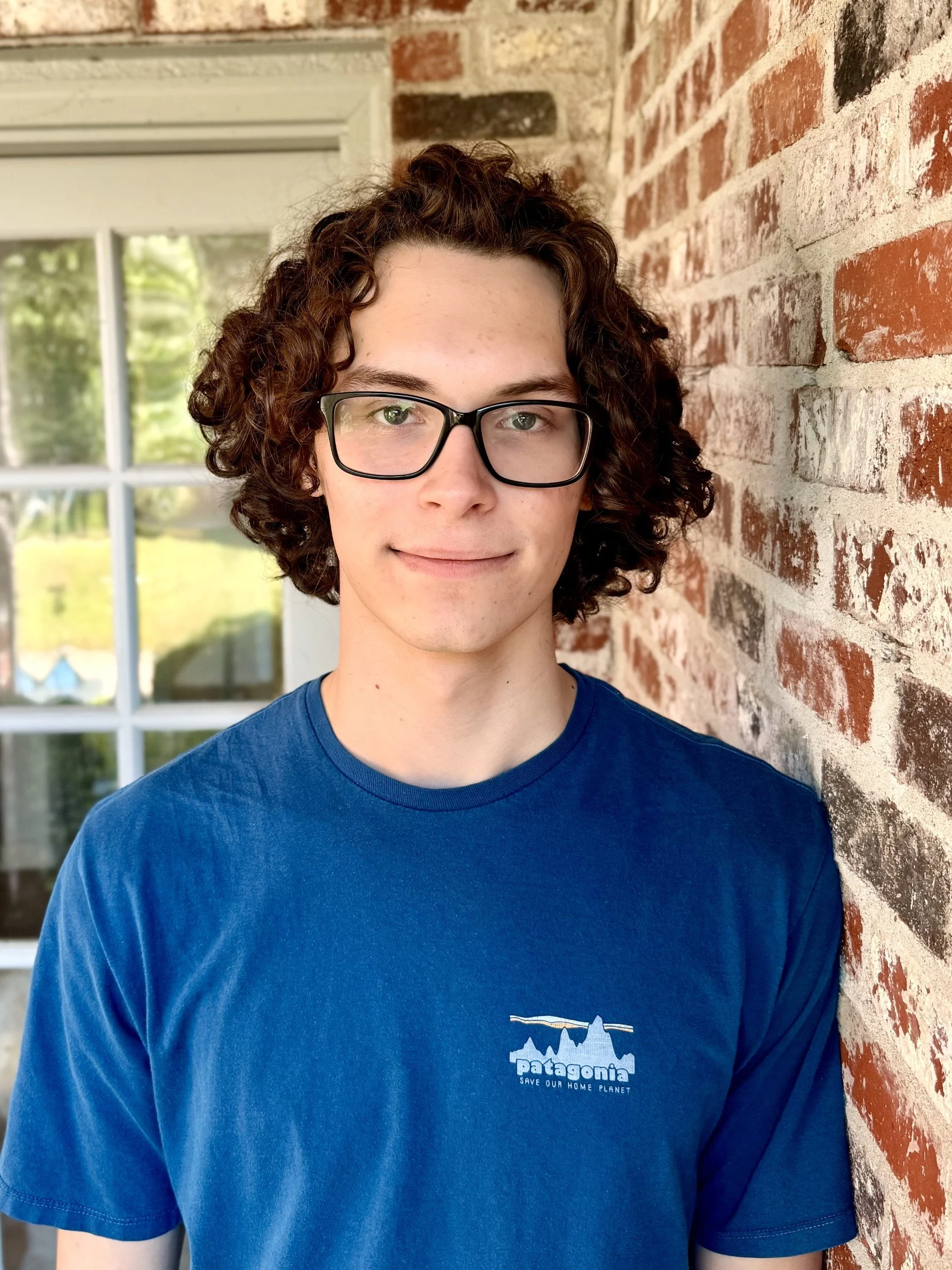 A boy with brown curly hair, glasses, and a blue shirt smiles at the camera