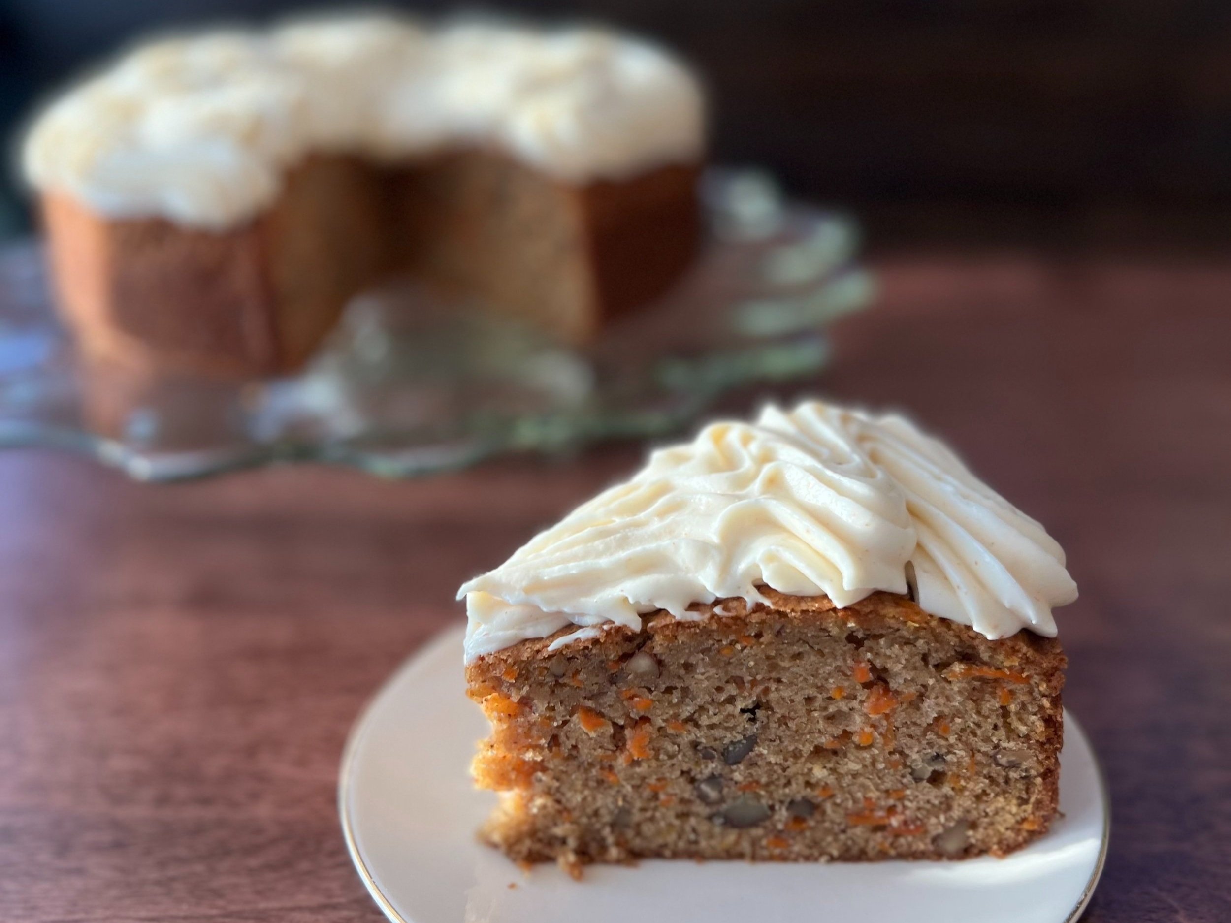 Slice of carrot cake with cream cheese frosting on a white plate, blurred whole cake in the background.