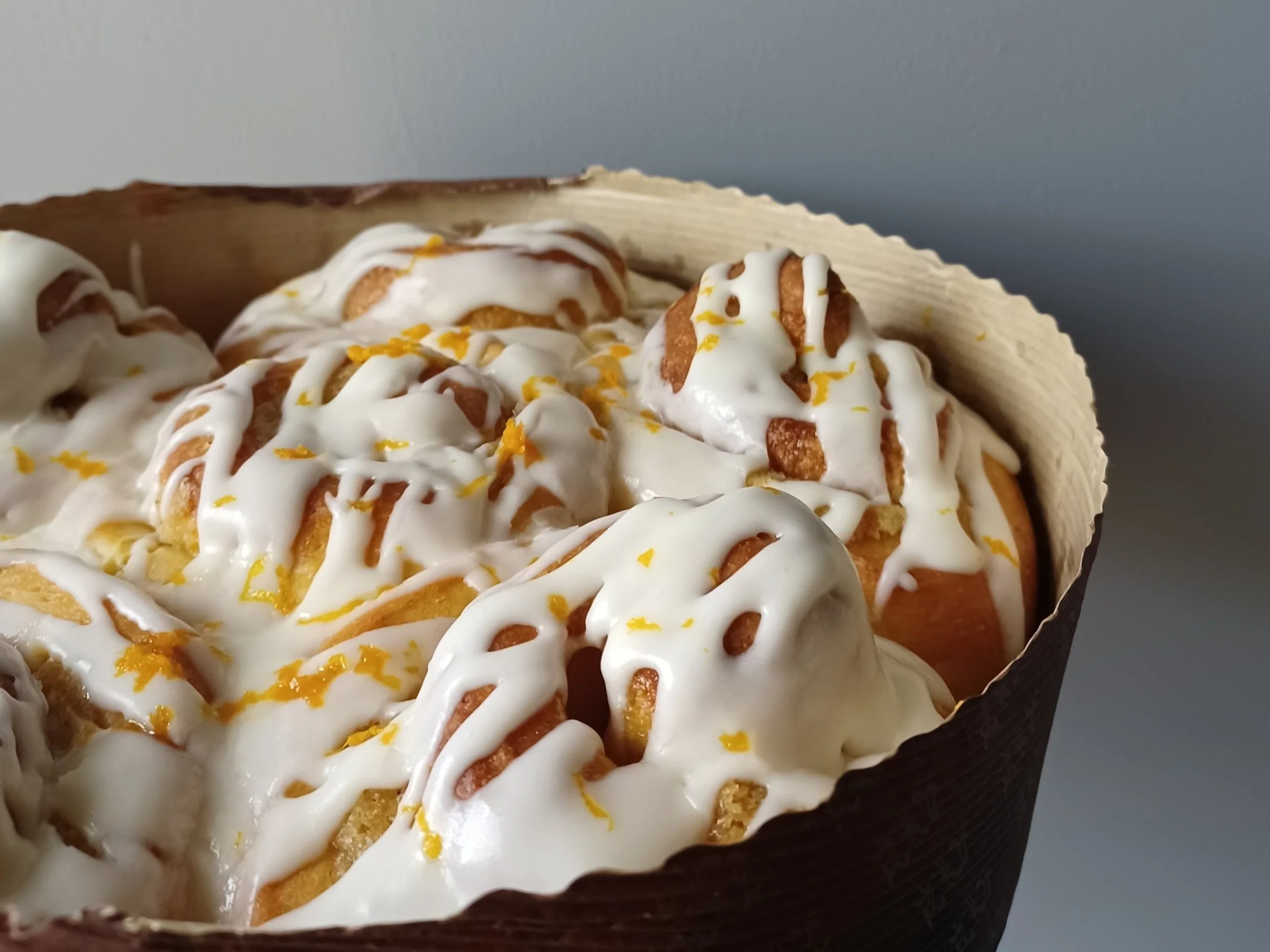 A close-up of cinnamon rolls topped with white icing and lemon zest in a black paper baking cup.