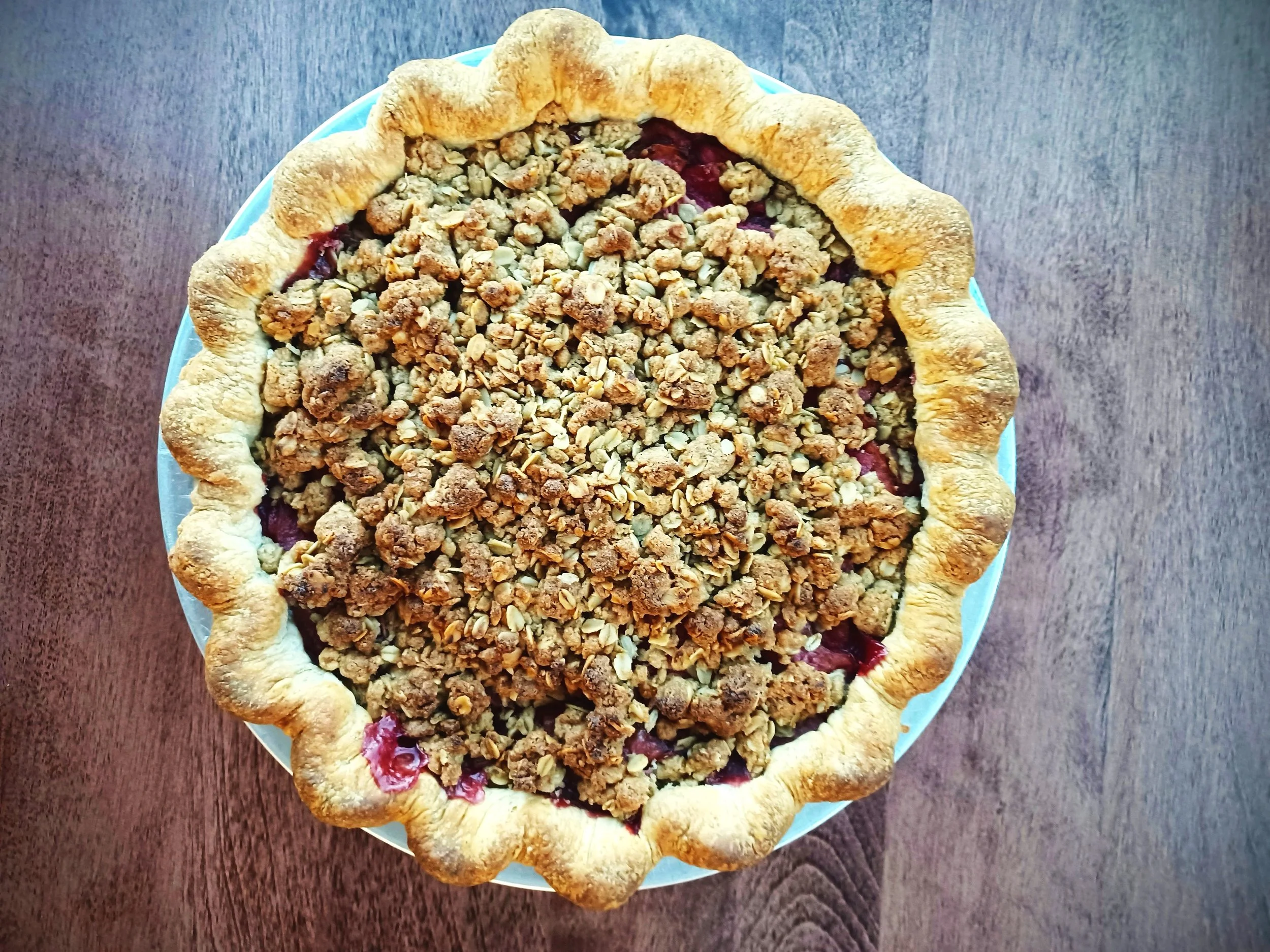 A pie with a golden crust and a crumbly topping, filled with a red fruit filling, placed on a blue plate on a wooden surface.
