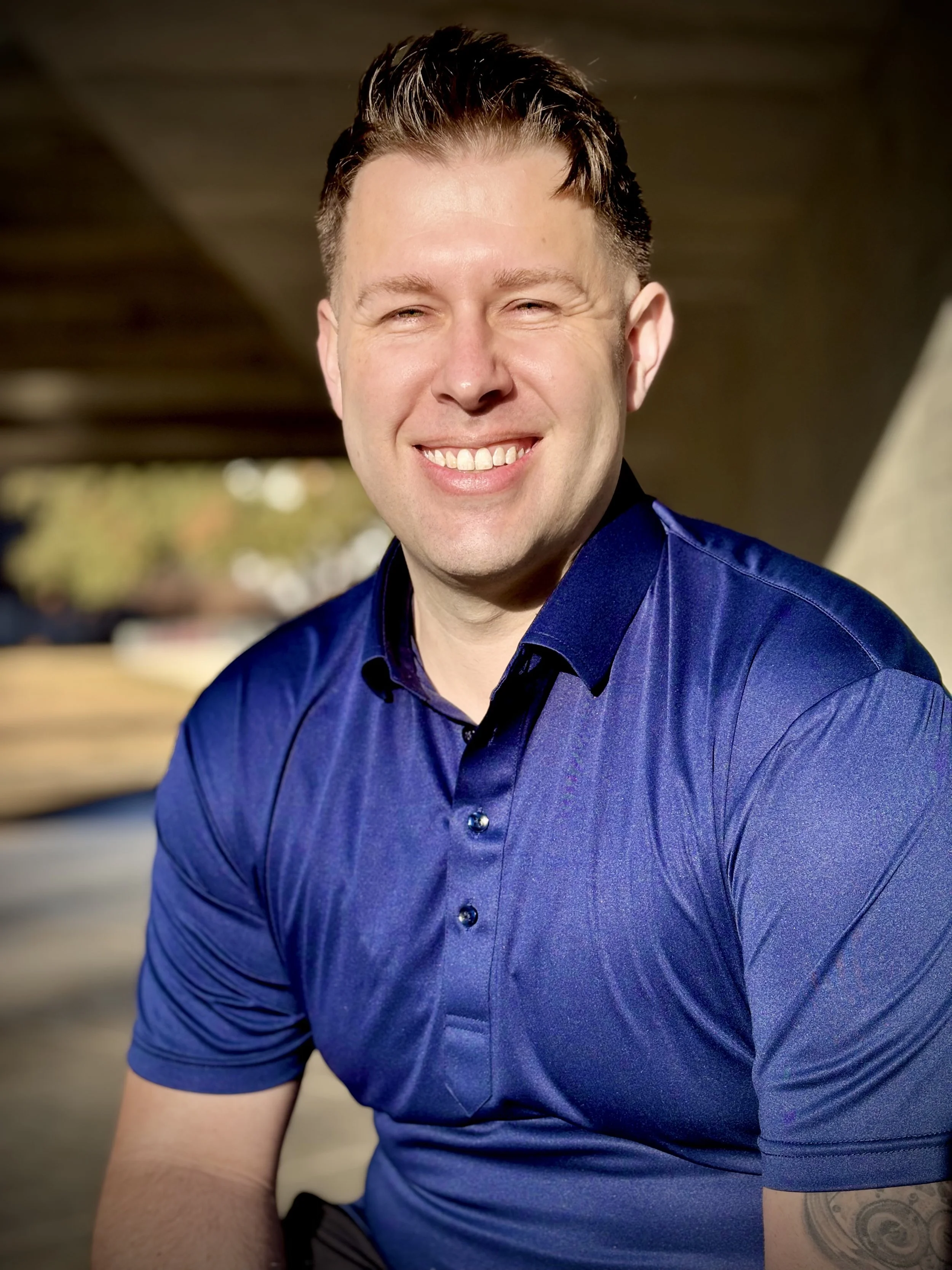 A smiling man with short, dark, styled hair, wearing a blue collared shirt, sitting outdoors with blurred background, showing a partial tattoo on his left arm.