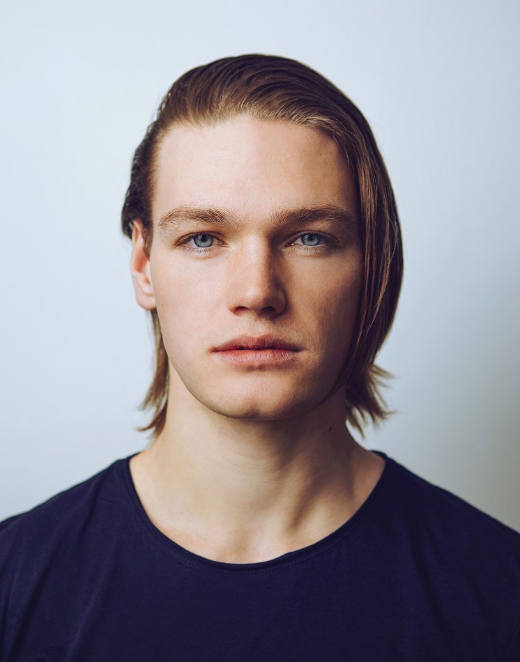 Close-up portrait of a young man with straight, shoulder-length light brown hair, blue eyes, and fair skin, wearing a black shirt, against a plain light background.