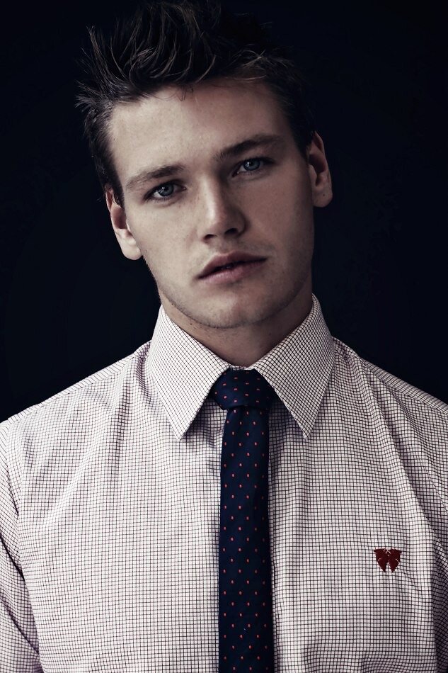 Young man wearing a checked dress shirt and dark patterned tie, with a small embroidered logo on the shirt's chest, against a dark background.