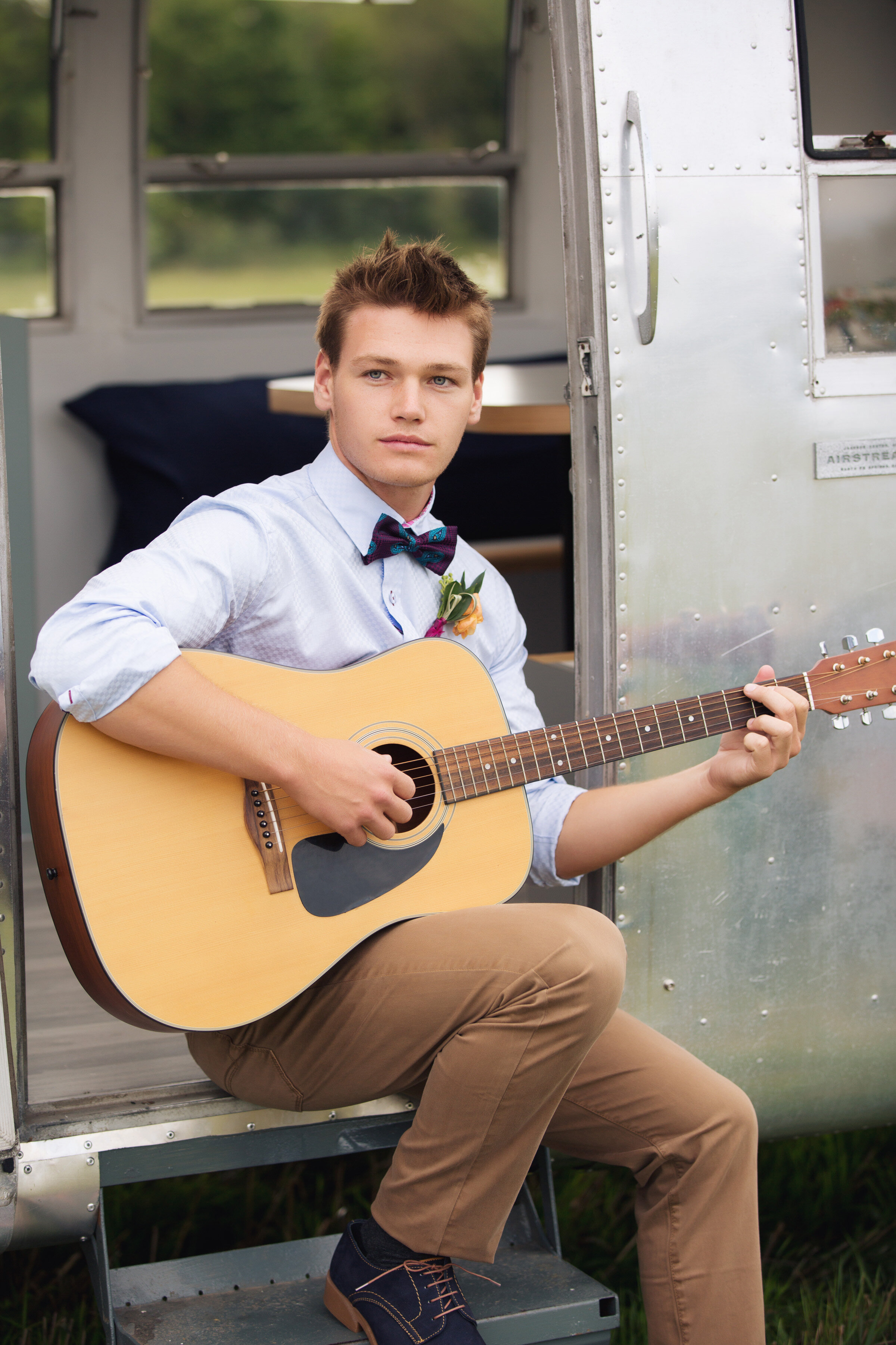 A young man with short brown hair sitting on the steps of a vintage trailer, playing an acoustic guitar outdoors, dressed in a light blue shirt, a purple bow tie, and tan pants.