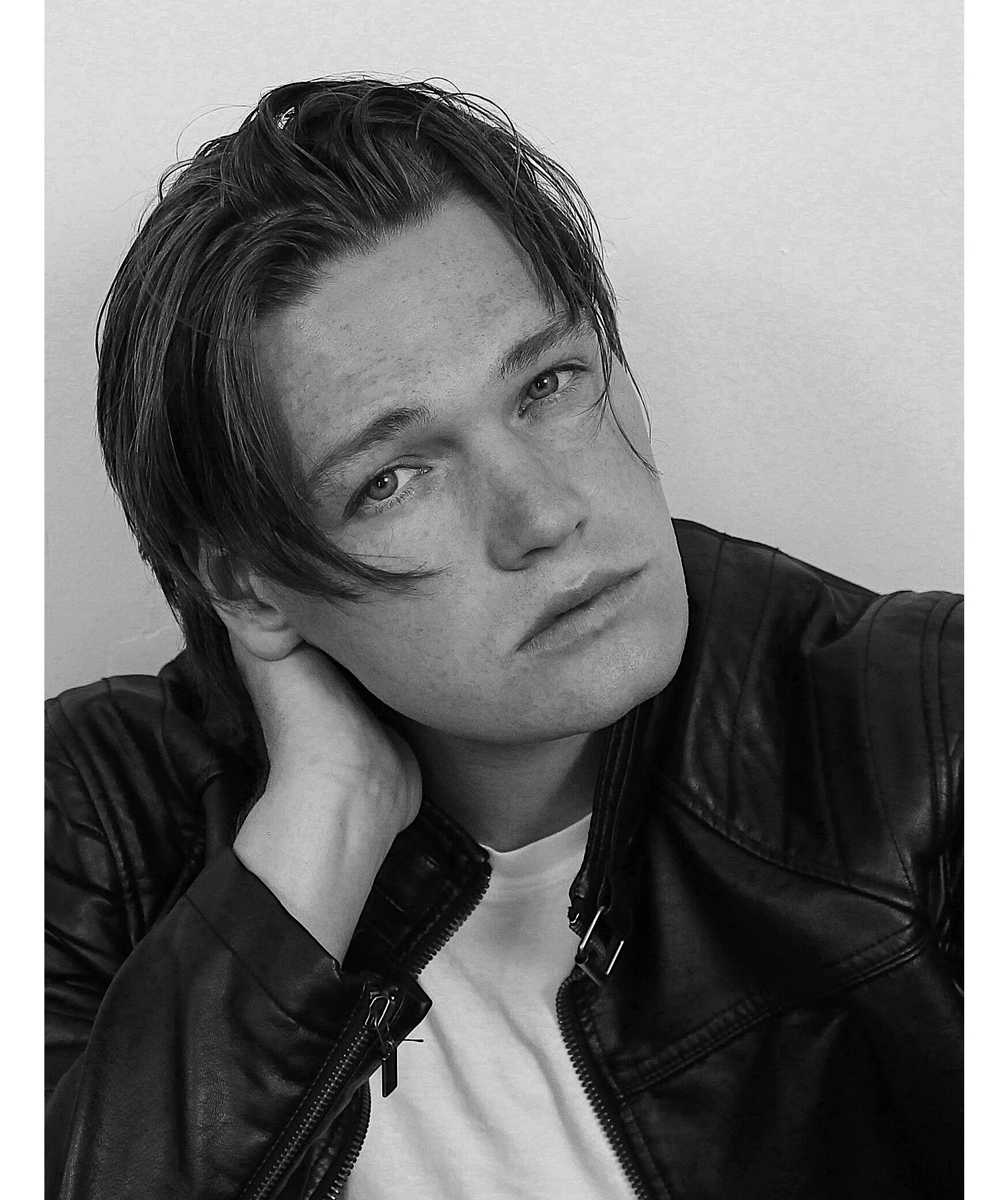Black and white photo of a young man with wet, side-parted hair, wearing a leather jacket and white T-shirt, resting his head on his hand and looking at the camera.
