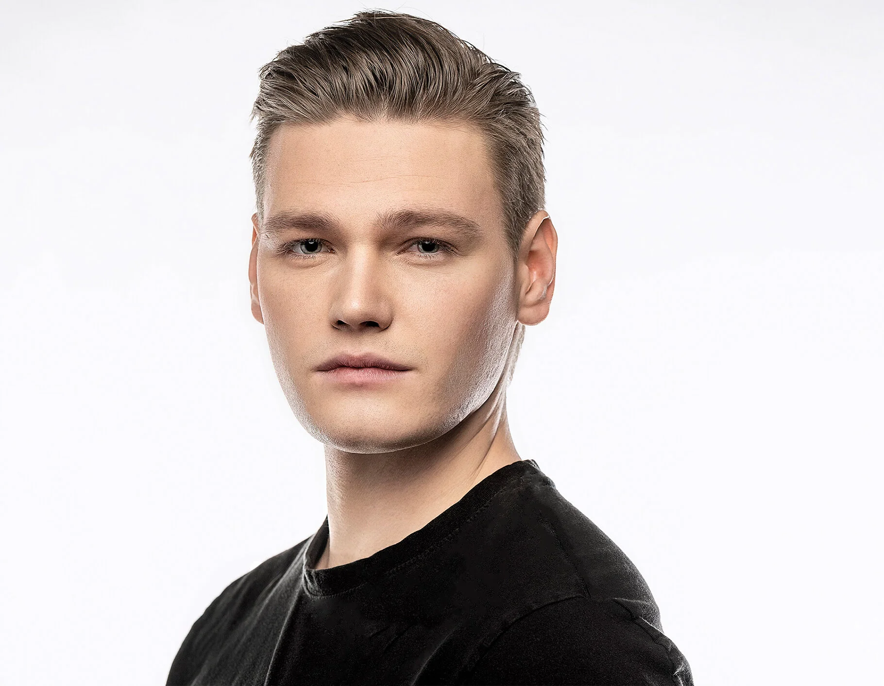 Portrait of a young man with slicked-back hair, fair skin, wearing a black shirt, against a plain white background.
