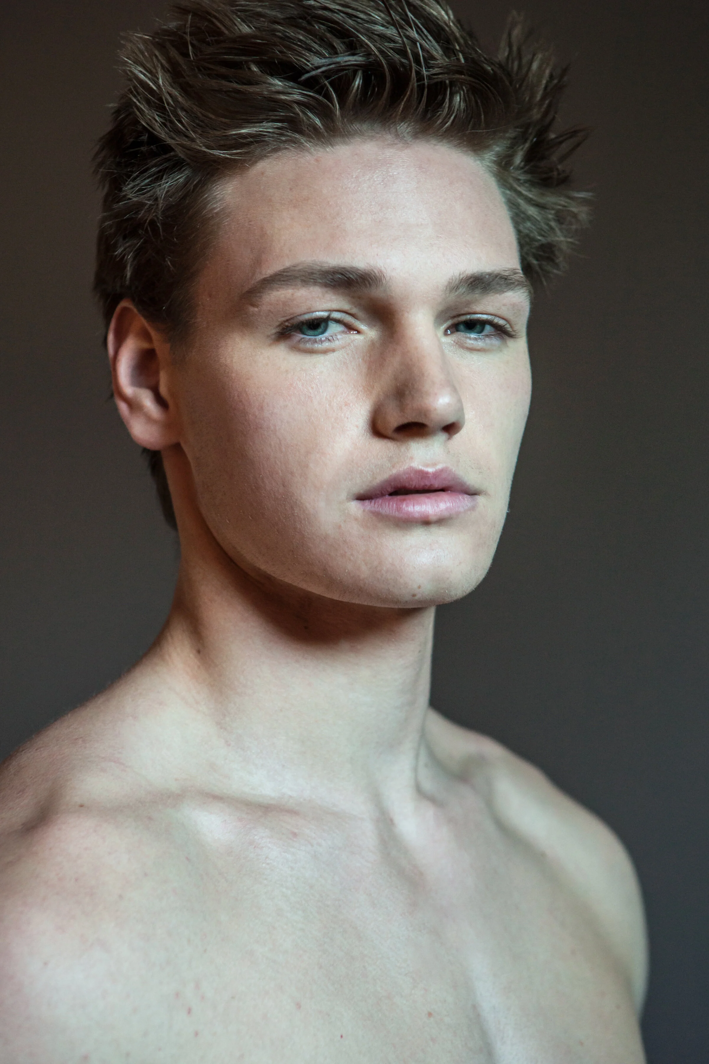 A close-up portrait of a young person with short, spiked brown hair, light skin, blue eyes, and a neutral expression, against a plain background.
