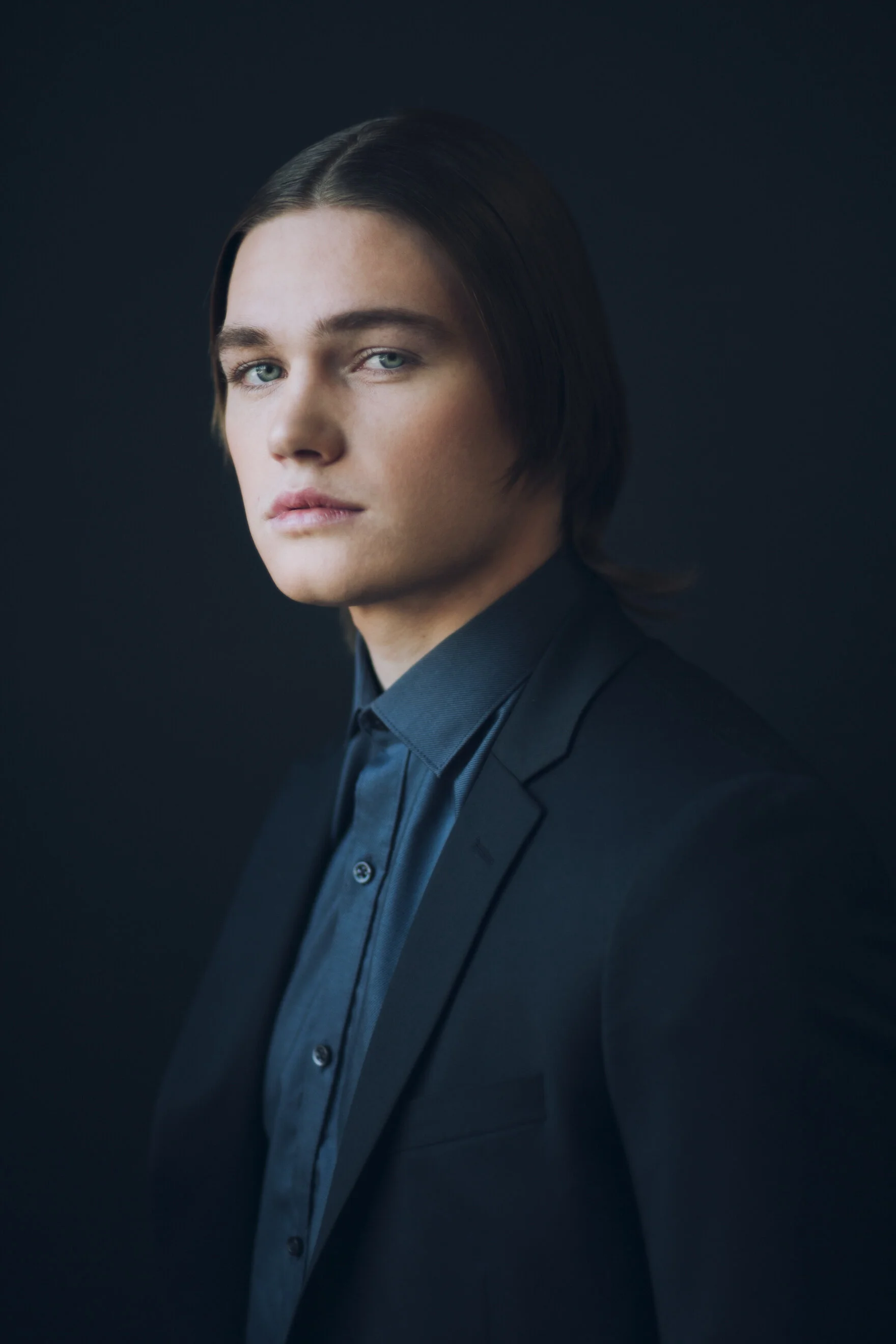 Portrait of a young man with dark hair, wearing a black suit and dark shirt, against a dark background.
