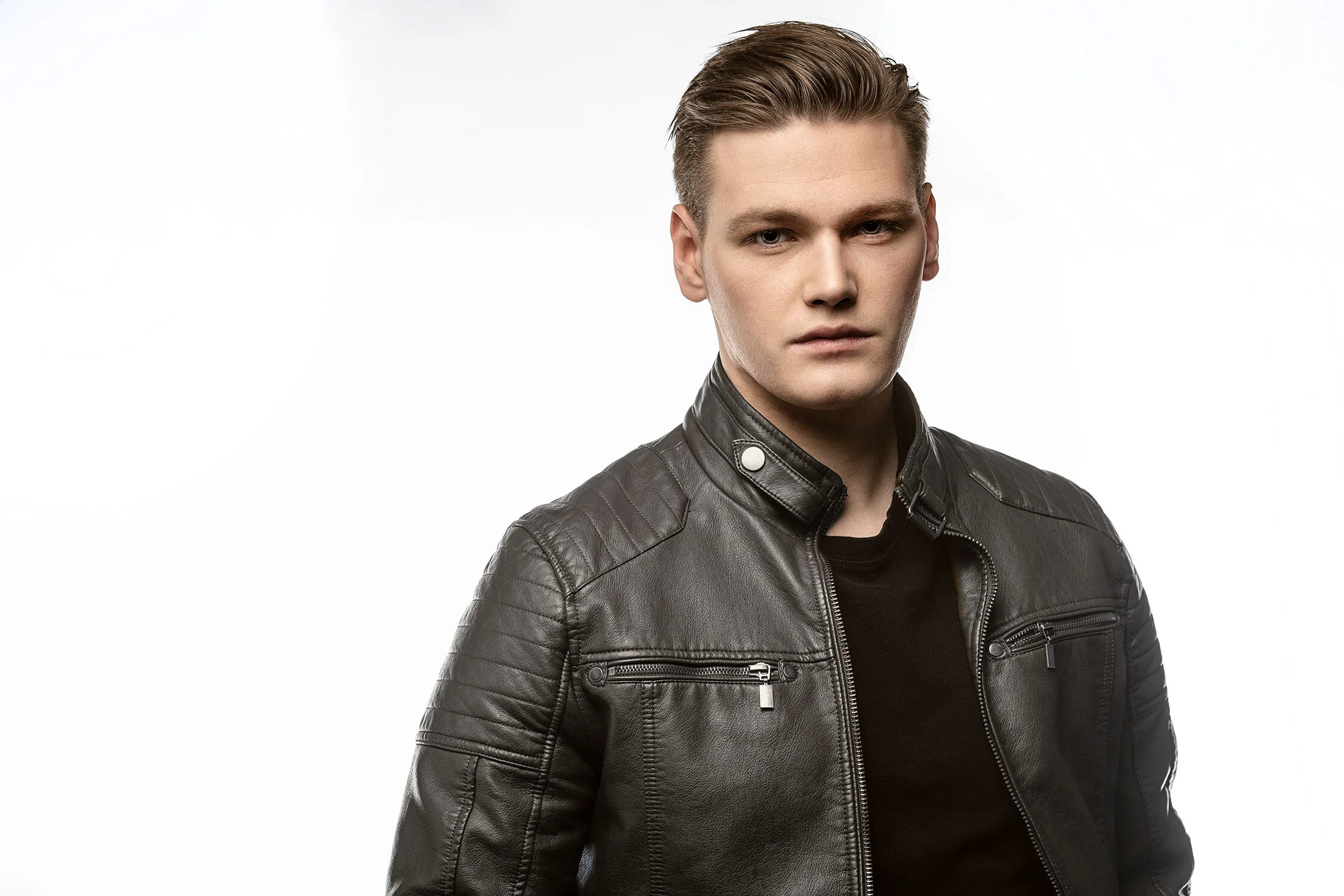 Young man with blonde hair wearing a black leather jacket, standing against a plain white background.